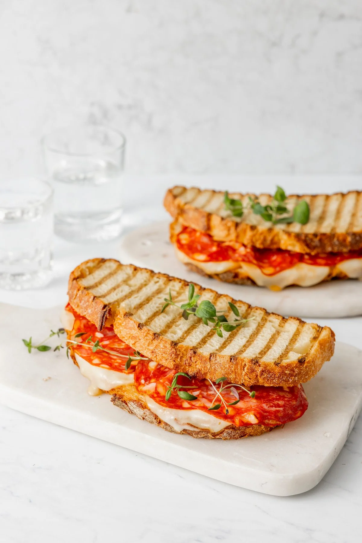 Close-up of a grilled toast with salami, melted cheese, and fresh herbs on a white marble serving board, with two glasses of water in the background.