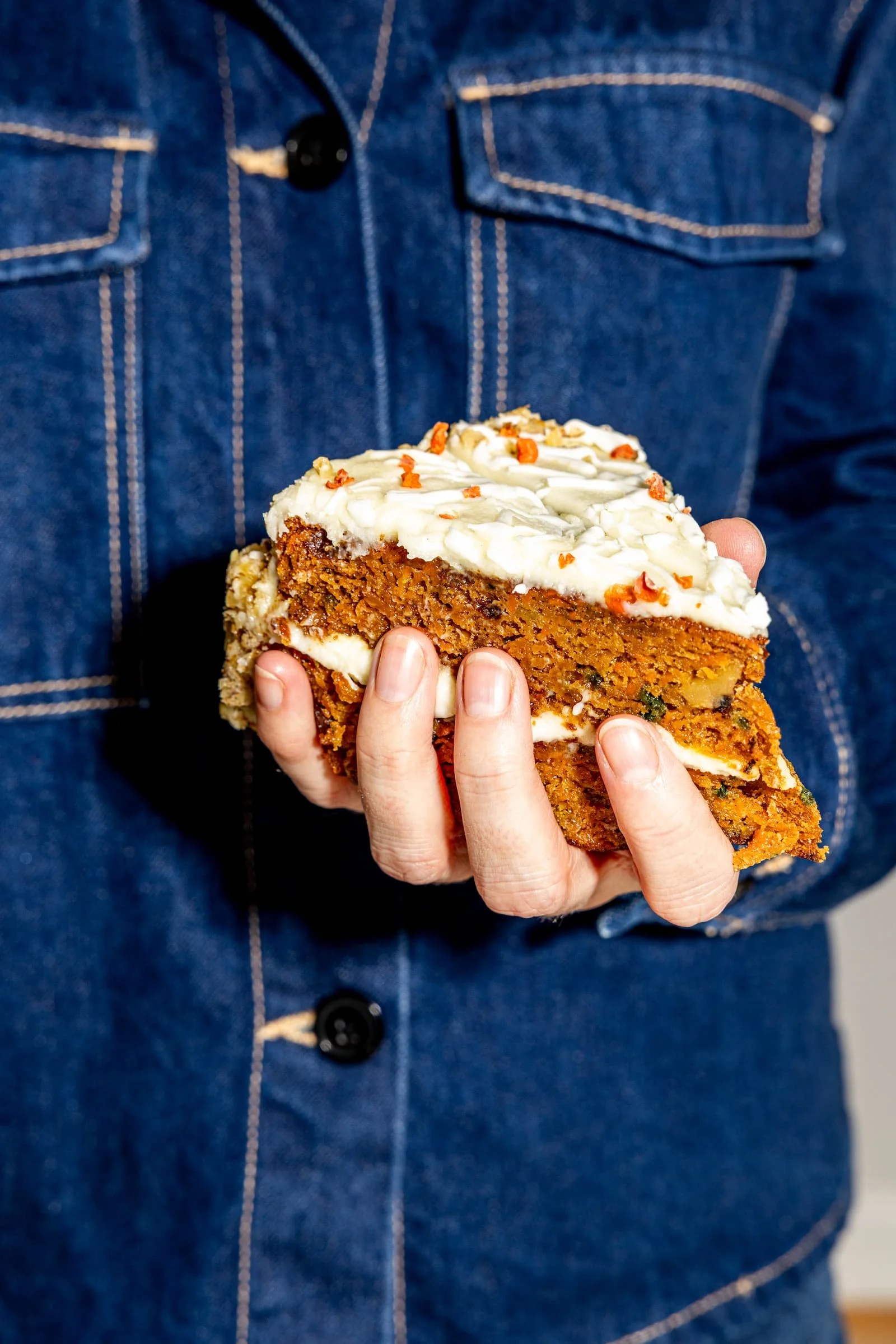 Person holding a slice of carrot cake with cream cheese frosting and orange sprinkles, wearing a denim jacket.
