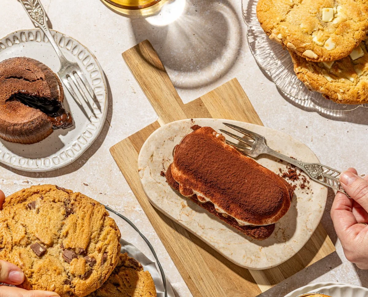 A variety of cookies placed on plates and a wooden serving board on a light-colored surface. There are chocolate chip cookies, a tiramisu cake, cookies, and chocolate fondant.
