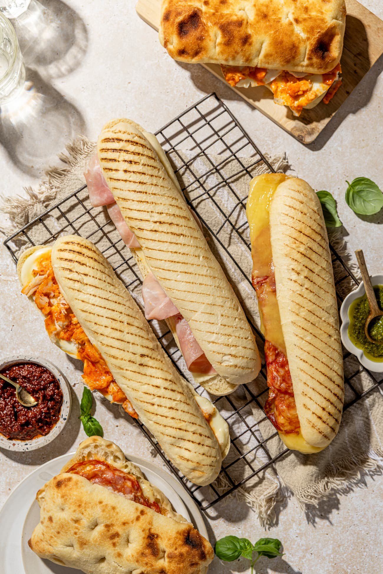 Three grilled panini sandwiches and one rolled flatbread with melted cheese and tomato sauce on a wire rack and a plate, with small bowls of pesto and tomato sauce nearby, on a light-colored surface.