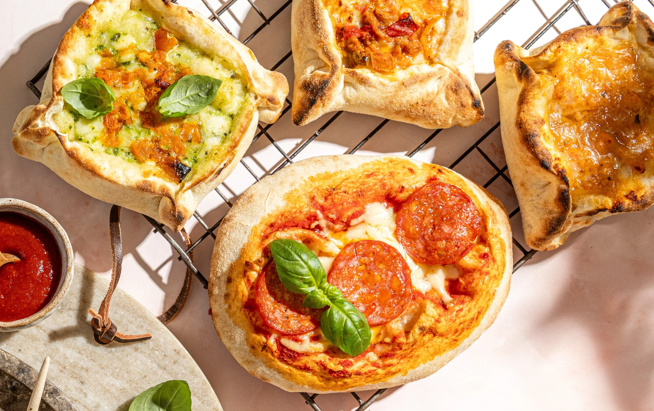 Assorted mini pizzas with toppings like pepperoni, cheese, basil, and vegetables on a cooling rack, with a side of marinara sauce.