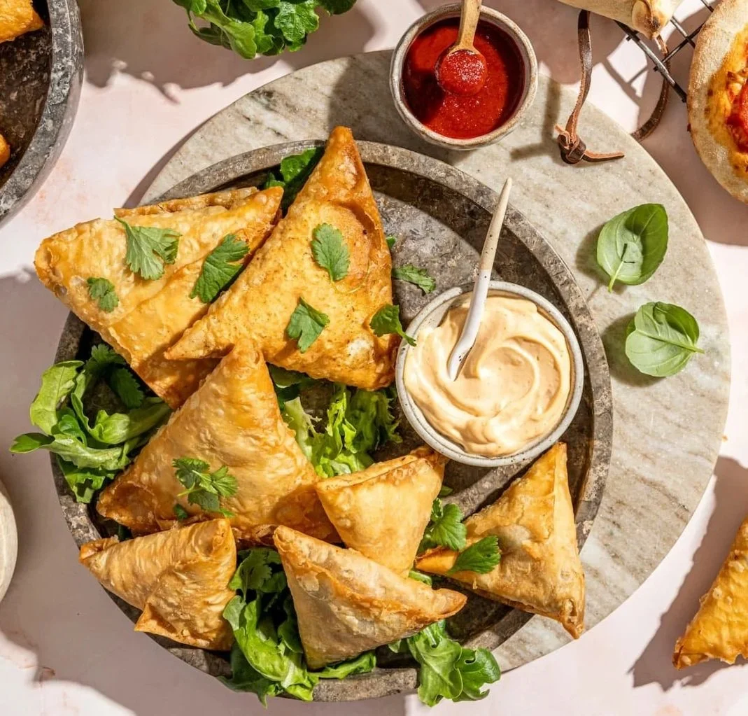 A platter of fried samosas garnished with cilantro, accompanied by a bowl of white dipping sauce, a small bowl of red chili sauce, and fresh green leaves around the plate.
