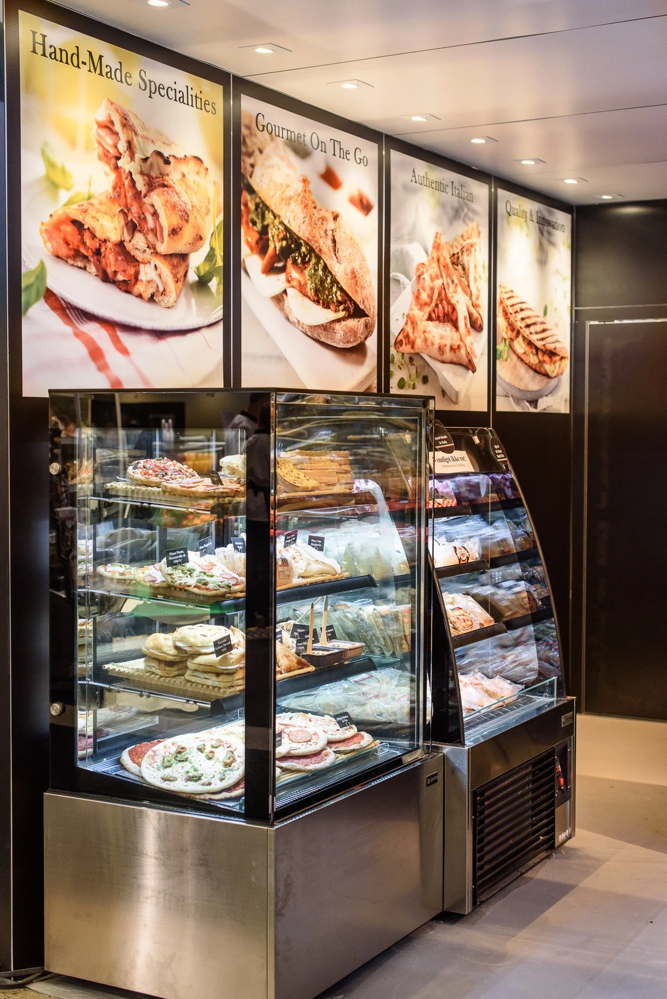 A display case of various pizzas and baked goods inside a pizzeria or bakery, with large photos of sandwiches and pizza toppings on the wall above.