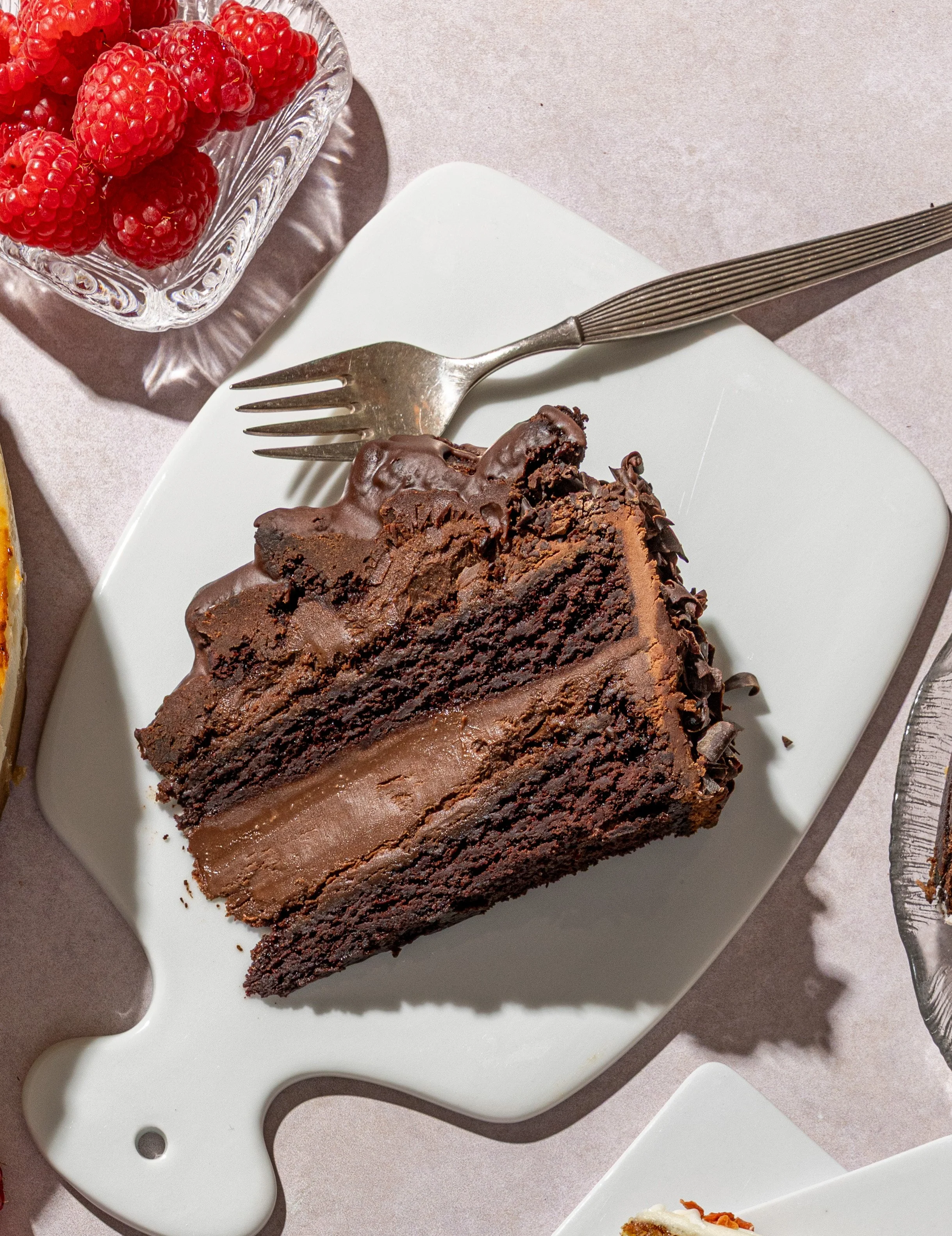 A slice of chocolate cake on a white serving board with a fork, a glass bowl of raspberries nearby, and other desserts partially visible around.