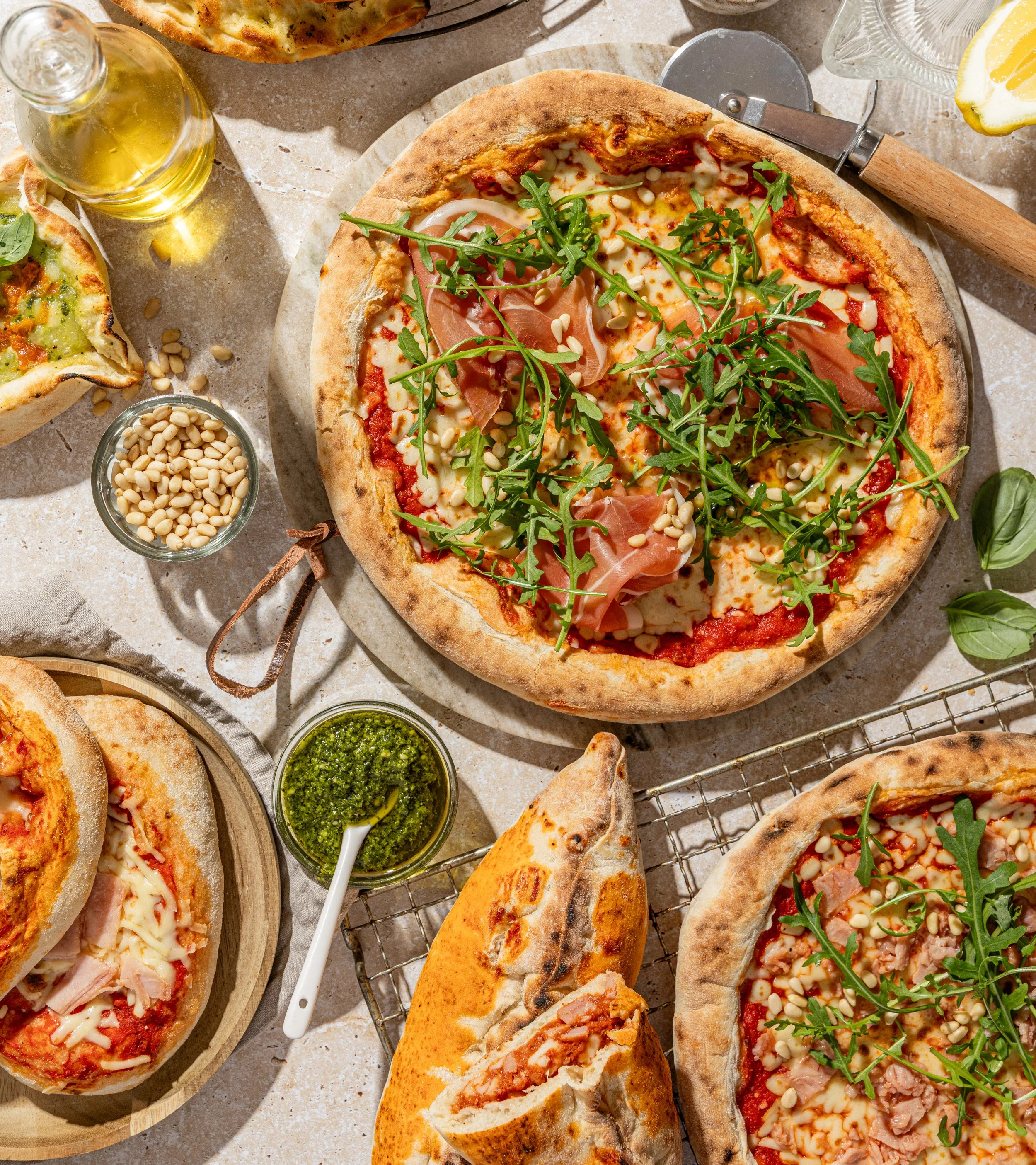 Assorted pizzas with toppings like arugula, prosciutto, and cheese, along with dipping sauces, lemon wedges, and olive oil on a light-colored surface.