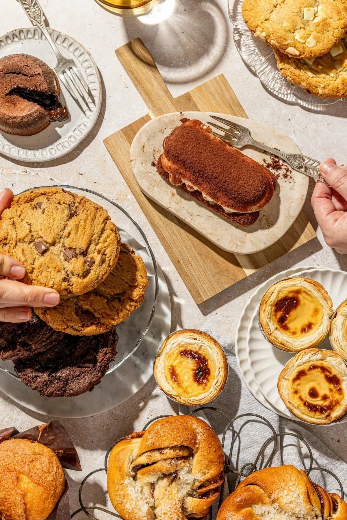 Assorted baked goods including cookies, pastel de nata, tiramisy, and handtwisted buns on plates and trays, with some hands reaching for treats.