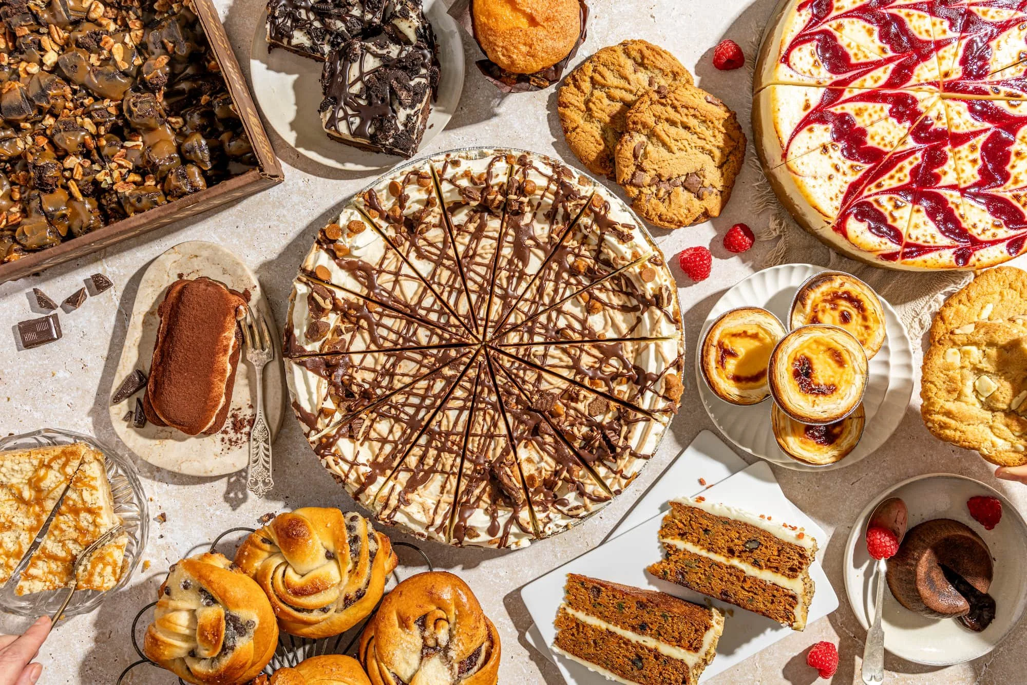 Assorted desserts including a chocolate chip cookie, a cheesecake drizzled with chocolate, a raspberry-topped cake, several pastries, and small cups of flan or custard, all arranged on a table with raspberries and small chocolates.