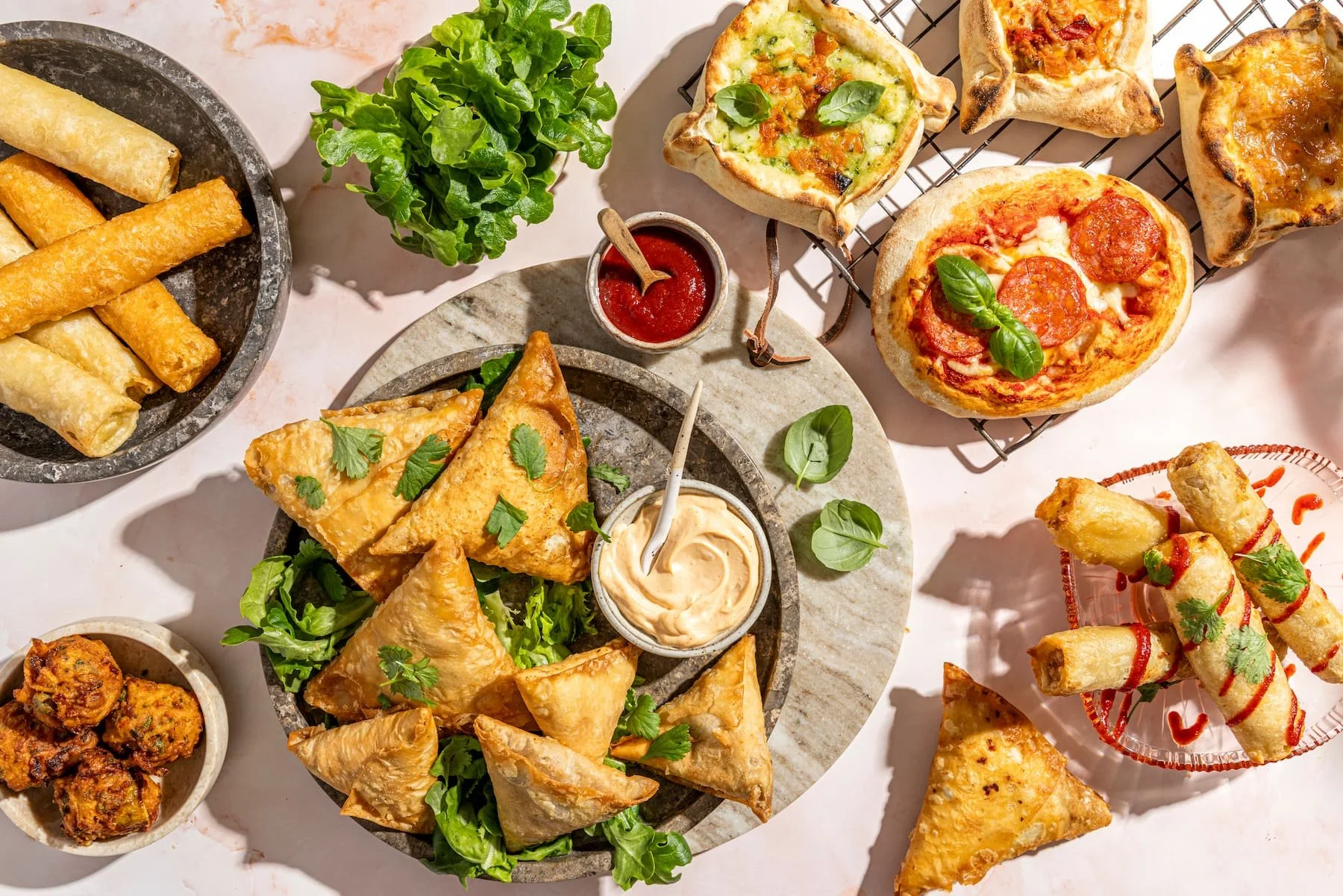 Assorted hot snacks on a table, including samosas, spring taquitos, pizza, and mini pizza bites, garnished with fresh herbs and served with dips.