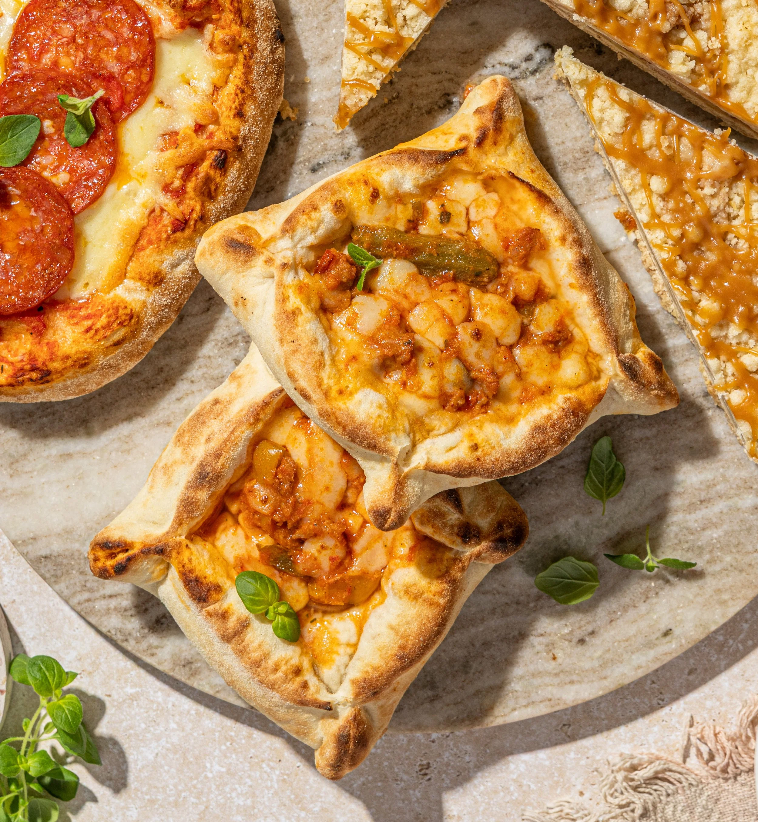 Two mini stuffed pizzas, one pepperoni pizza, and two slices of apple pie on a round beige plate garnished with small green leaves.