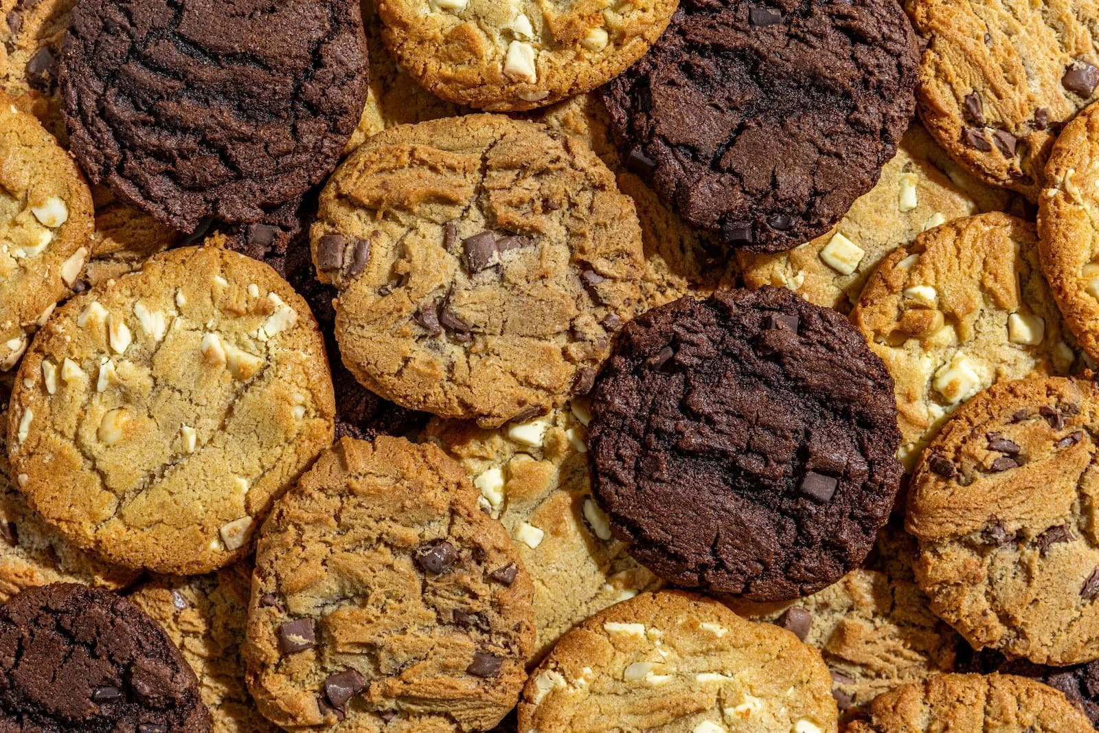 A close-up view of various chocolate chip cookies, including some with white chocolate and nuts, arranged in a pile.