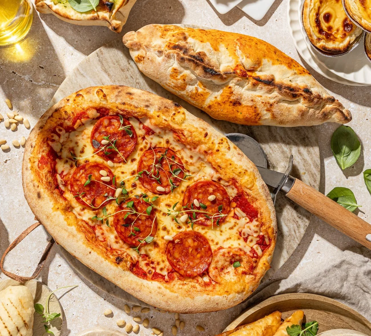 A pizza with pepperoni, cheese, and herbs, a calzone, and a small plate of pastel de nata desserts on a marble surface.