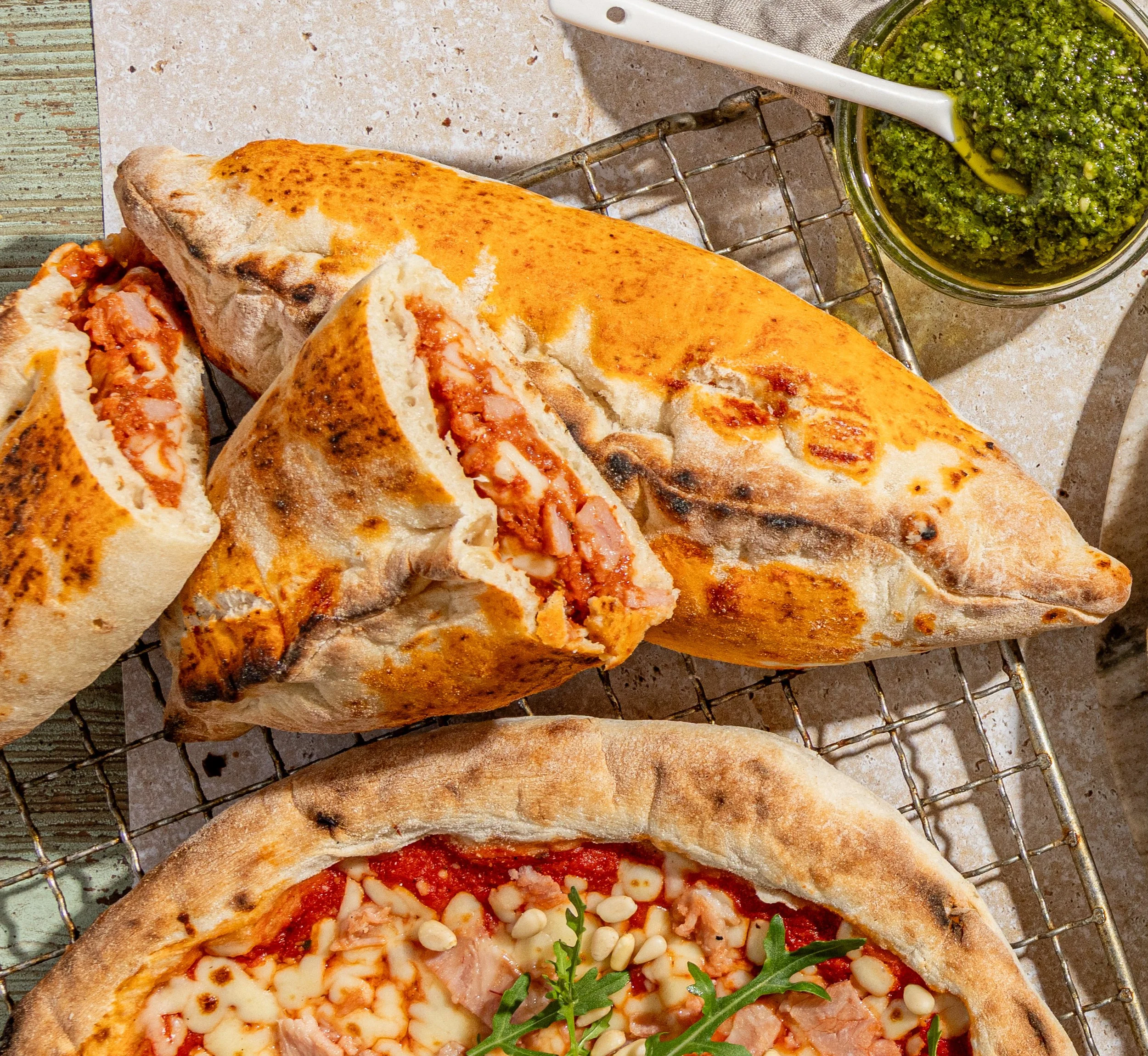 Two pizza calzones with melted cheese and tomato sauce, one with visible ham filling, on a cooling rack, with a green pesto sauce in a small jar nearby.