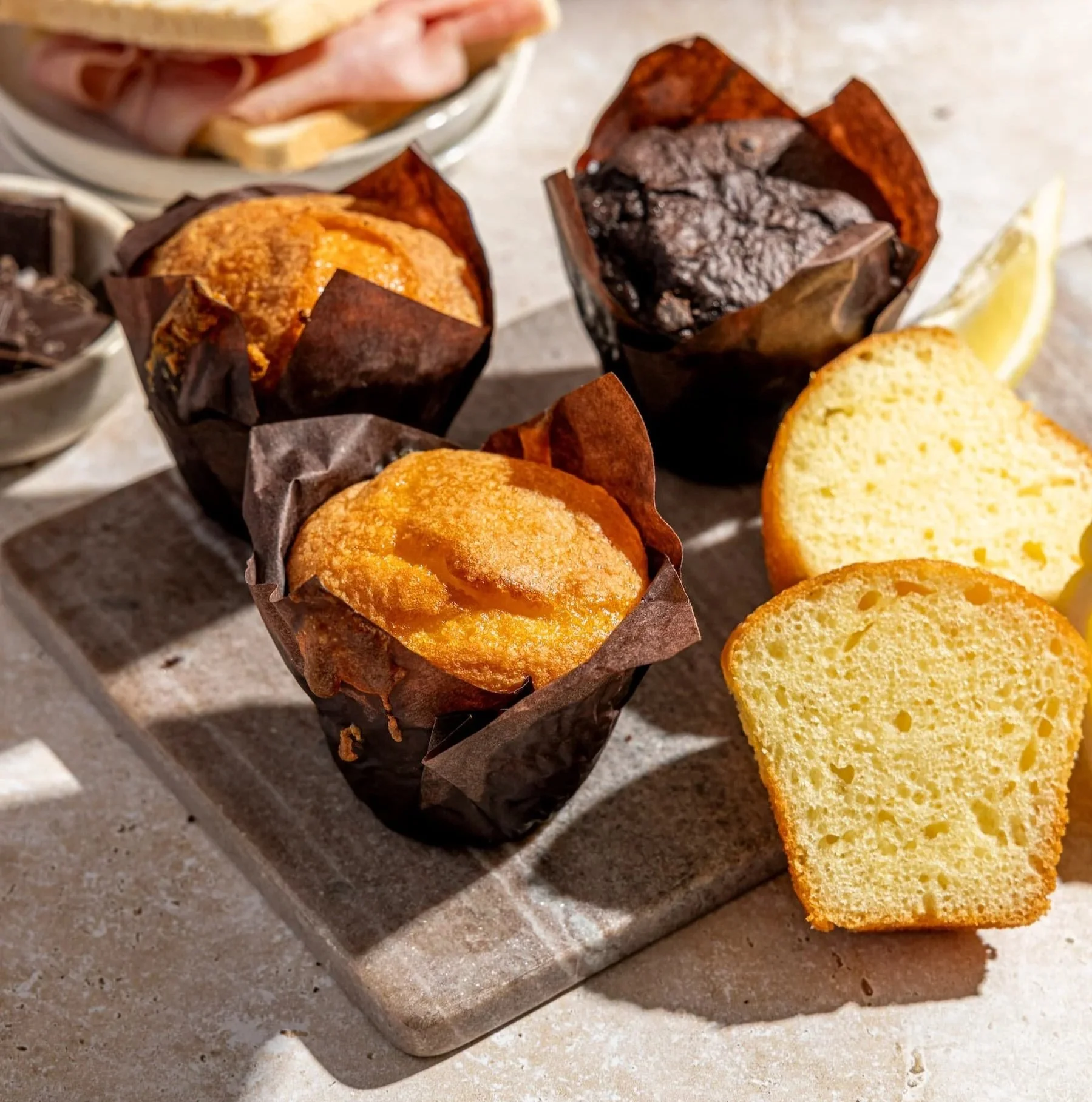 Three muffins in paper wrappers, one chocolate and two lemon, on a gray platter with slices of yellow pound cake and lemon wedges in the background.
