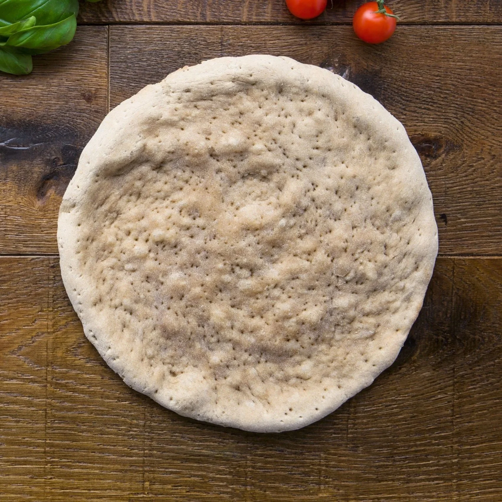 Unbaked pizza crust on a wooden surface with cherry tomatoes and basil in the background.