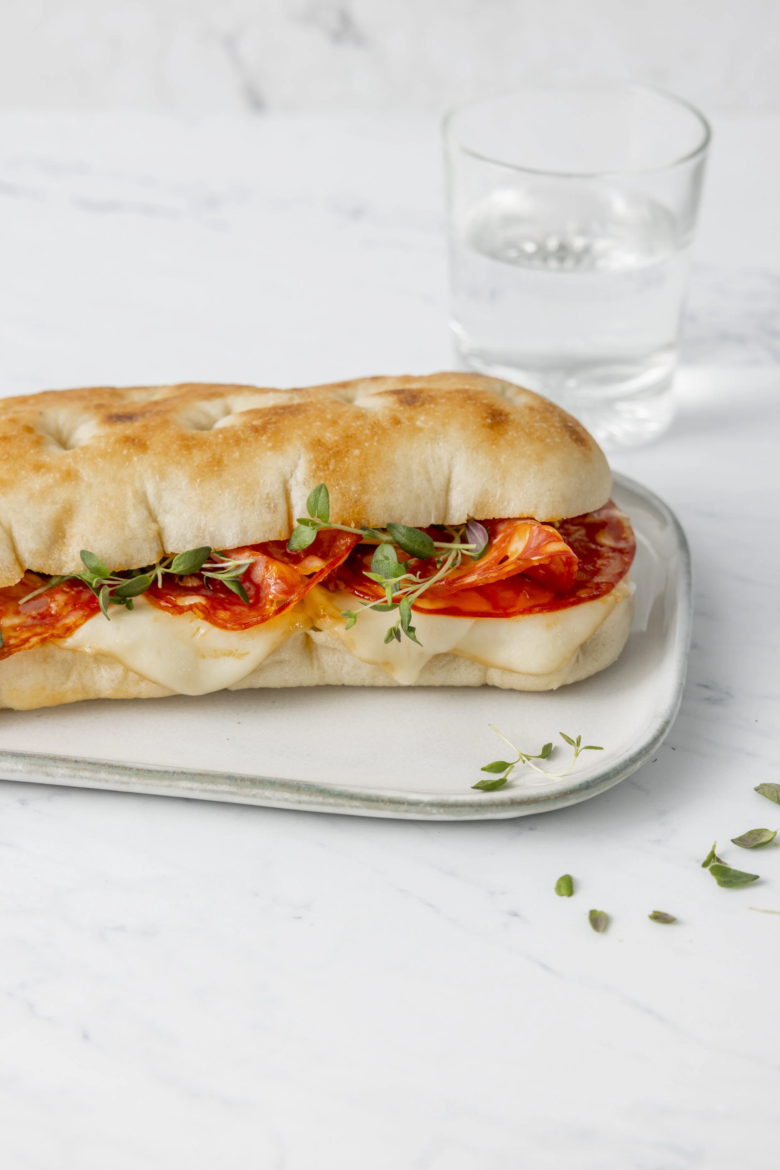 An artisan panini with melted cheese and salami, and fresh herbs on a plate with a glass of water in the background.