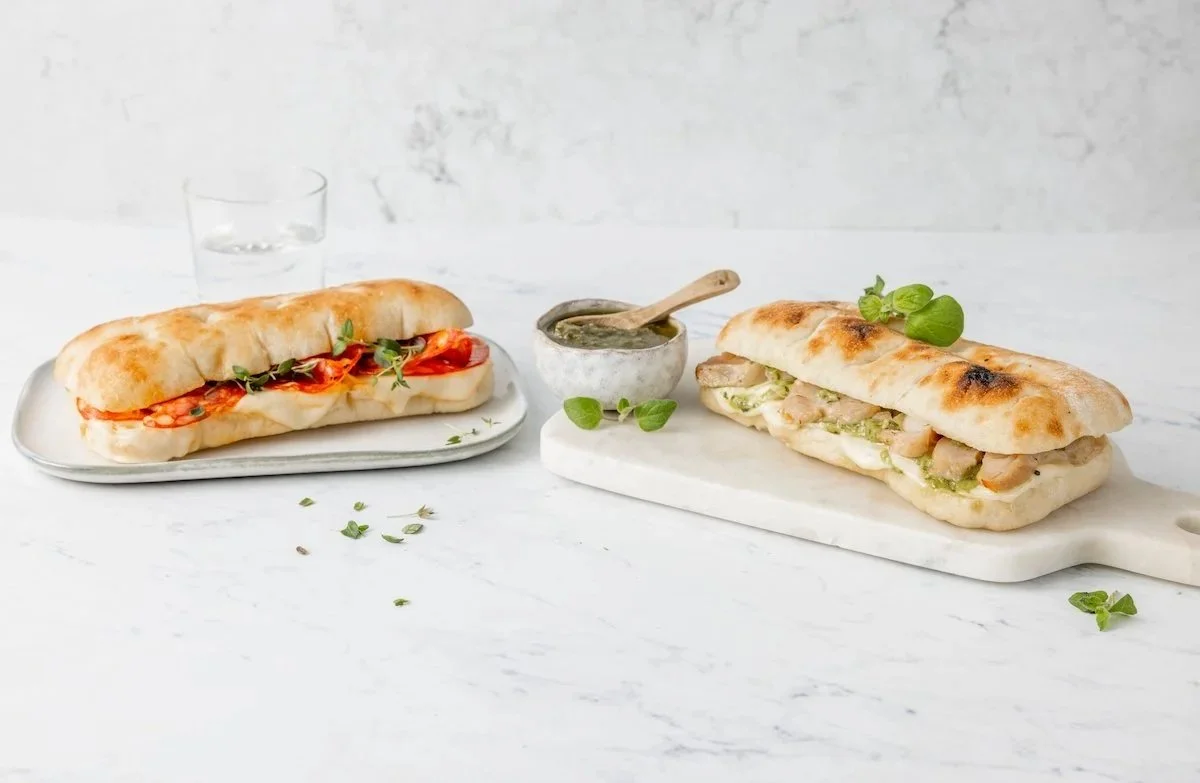 Two artisan paninis on a white marble surface, with a small bowl of dipping sauce in between, on a light background.