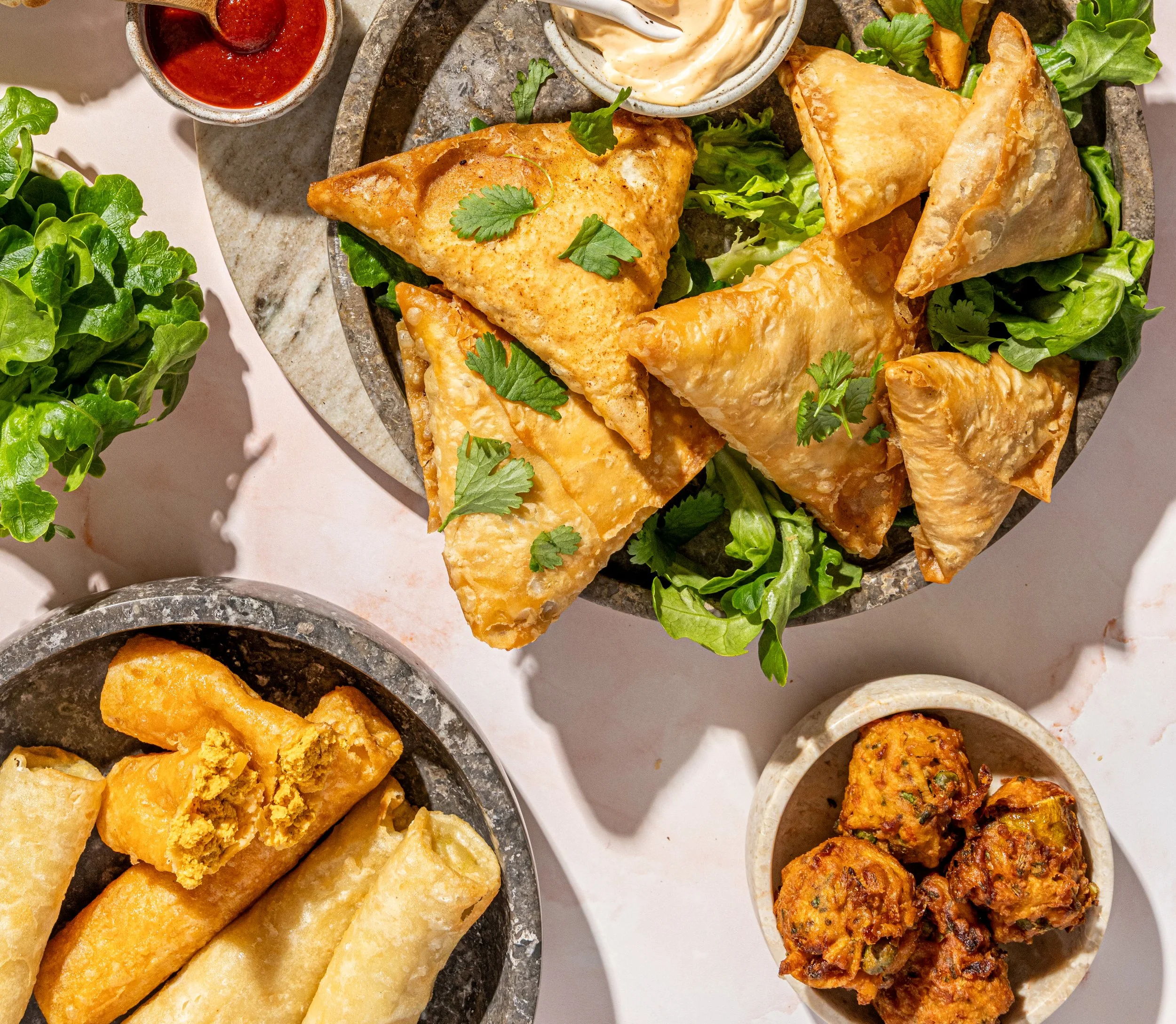 A top-down view of a platter of samosas garnished with cilantro, served with a bowl of yogurt and a small container of red sauce. Nearby, there are taquitos and spiced potato balls on separate plates, with a small plant of green lettuce.