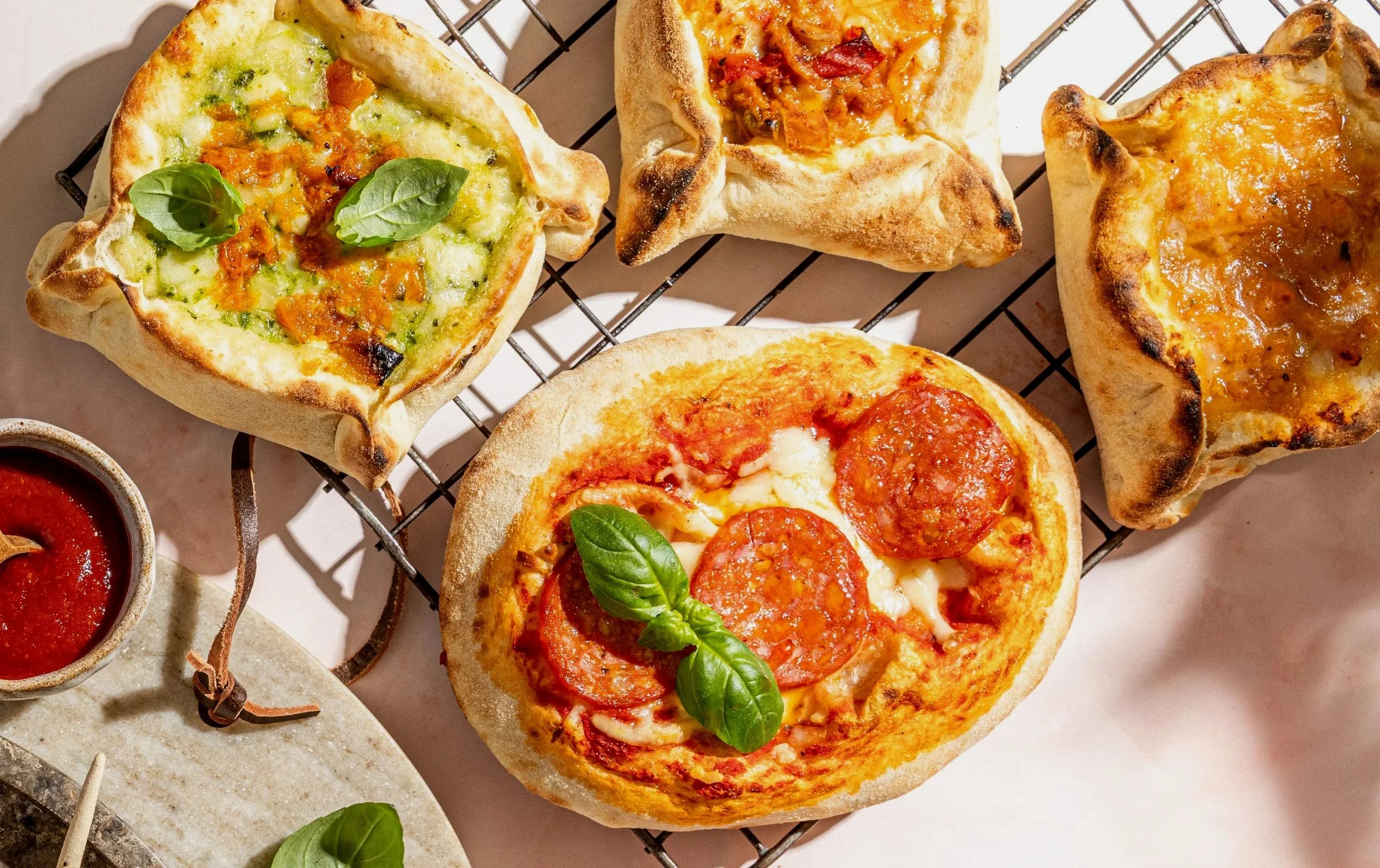 Assorted mini pizzas with toppings like pepperoni, cheese, and vegetables, on a wire cooling rack, accompanied by a small bowl of tomato sauce.