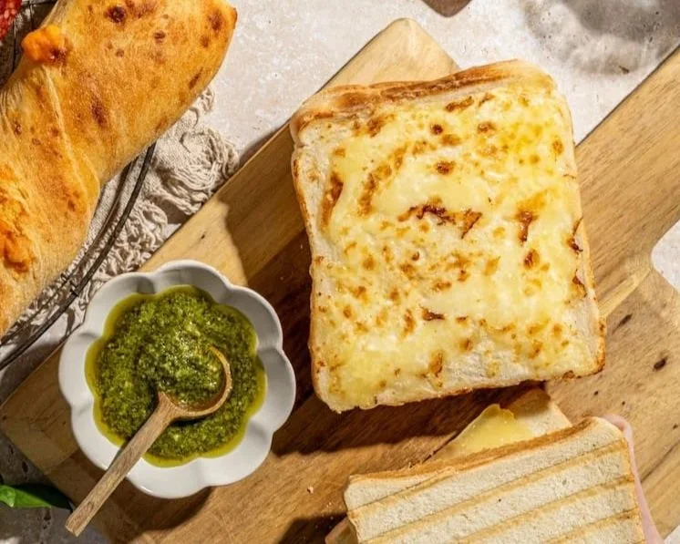 A piece of toasted bread topped with melted cheese, a small bowl of green pesto sauce nearby, and parts of other toasted bread in the background on a wooden cutting board.