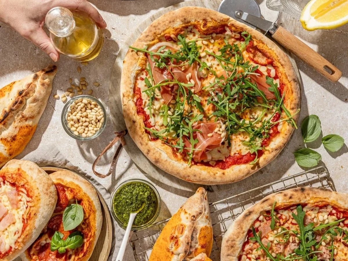 Assorted pizzas with arugula and pine nuts, accompanied by a bottle of olive oil, basil, and green pesto.