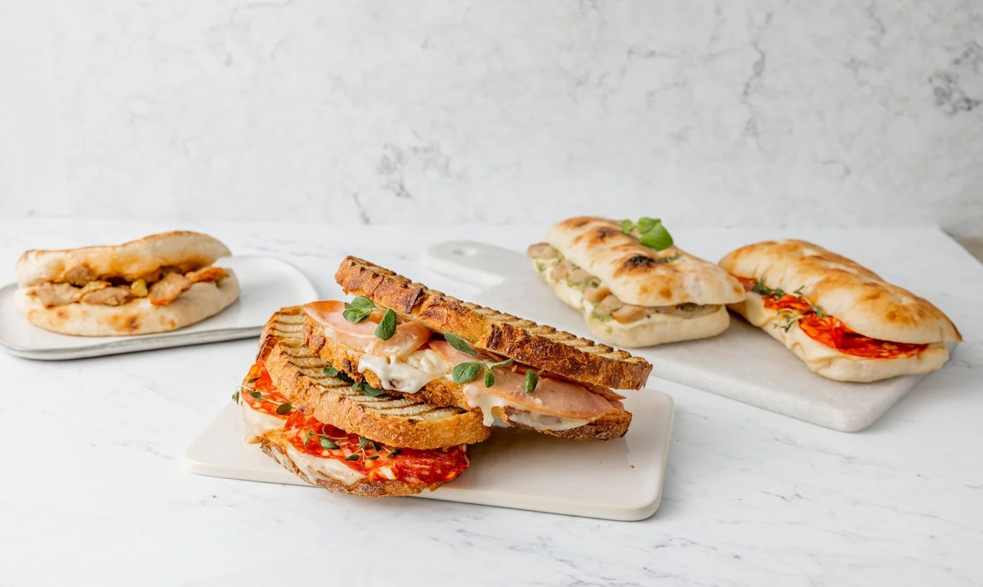 Assorted sandwiches and flatbreads on marble surfaces, with one sandwich in the foreground filled with sliced tomatoes, cured meats, herbs, and mayonnaise, and others in the background with various fillings.