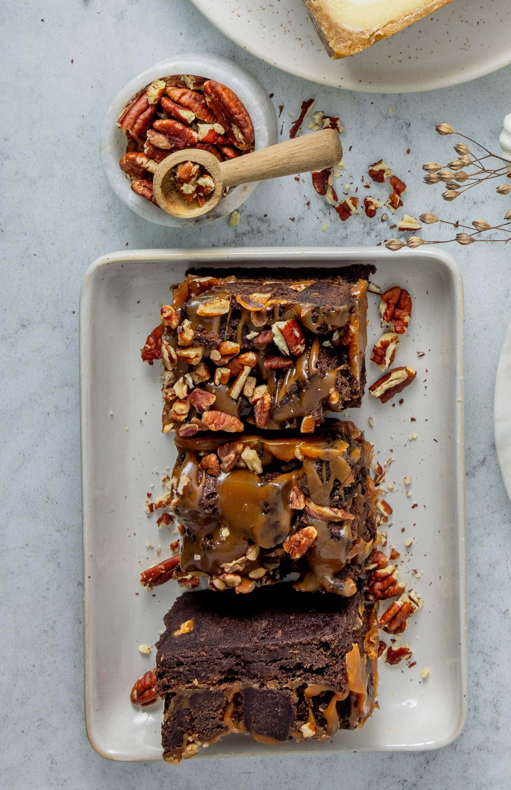 Three chocolate brownies topped with caramel and chopped pecans on a white rectangular plate, with a small bowl of pecans and a scoop in the background.