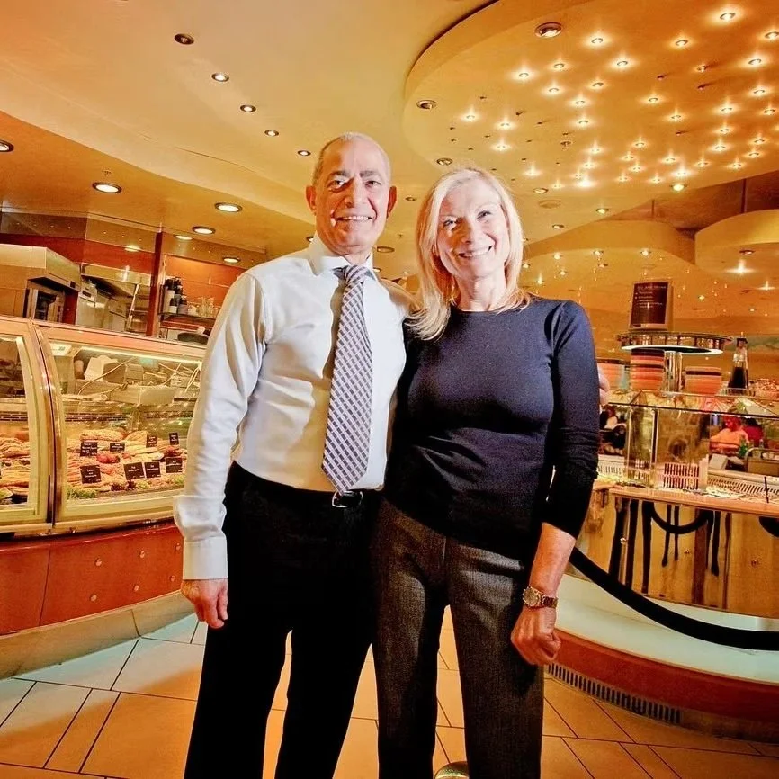 A man and woman smiling and standing close together inside a restaurant or cafe, with a display case of desserts behind them and a warm, yellowish ceiling with many small lights.