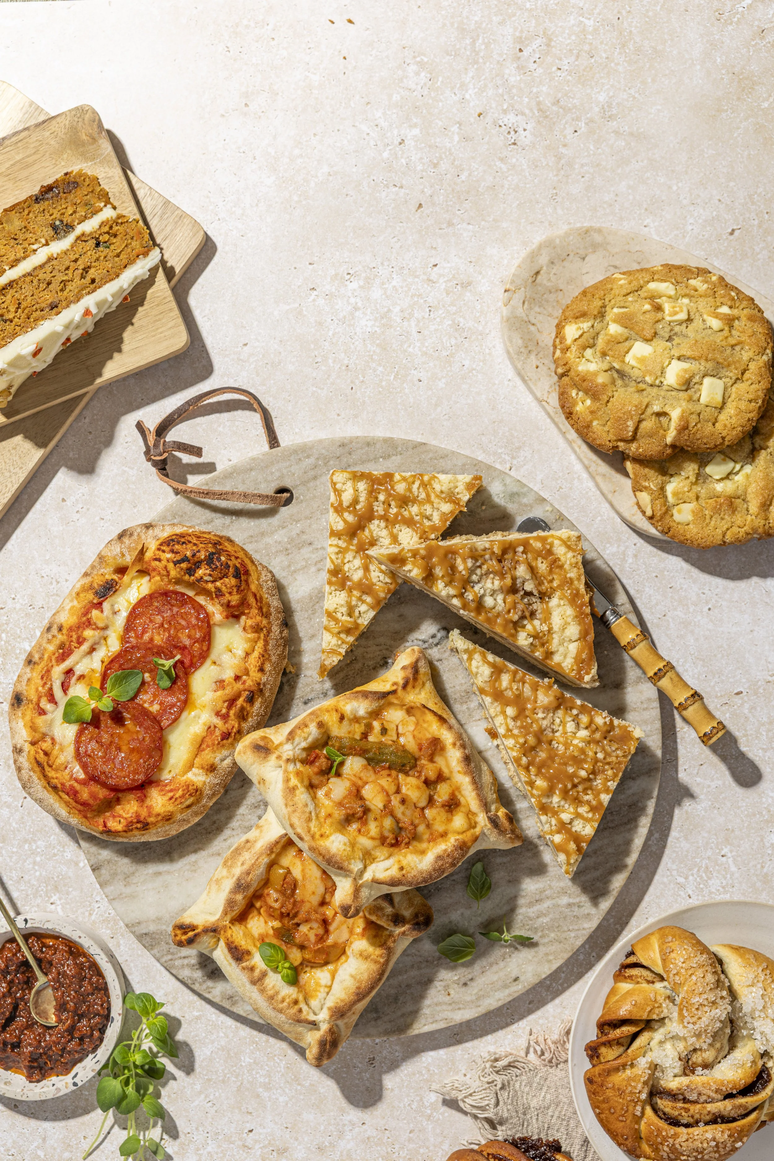 Assorted bakery products, like cookies and cakes, plus pizza and piazza on a light surface.
