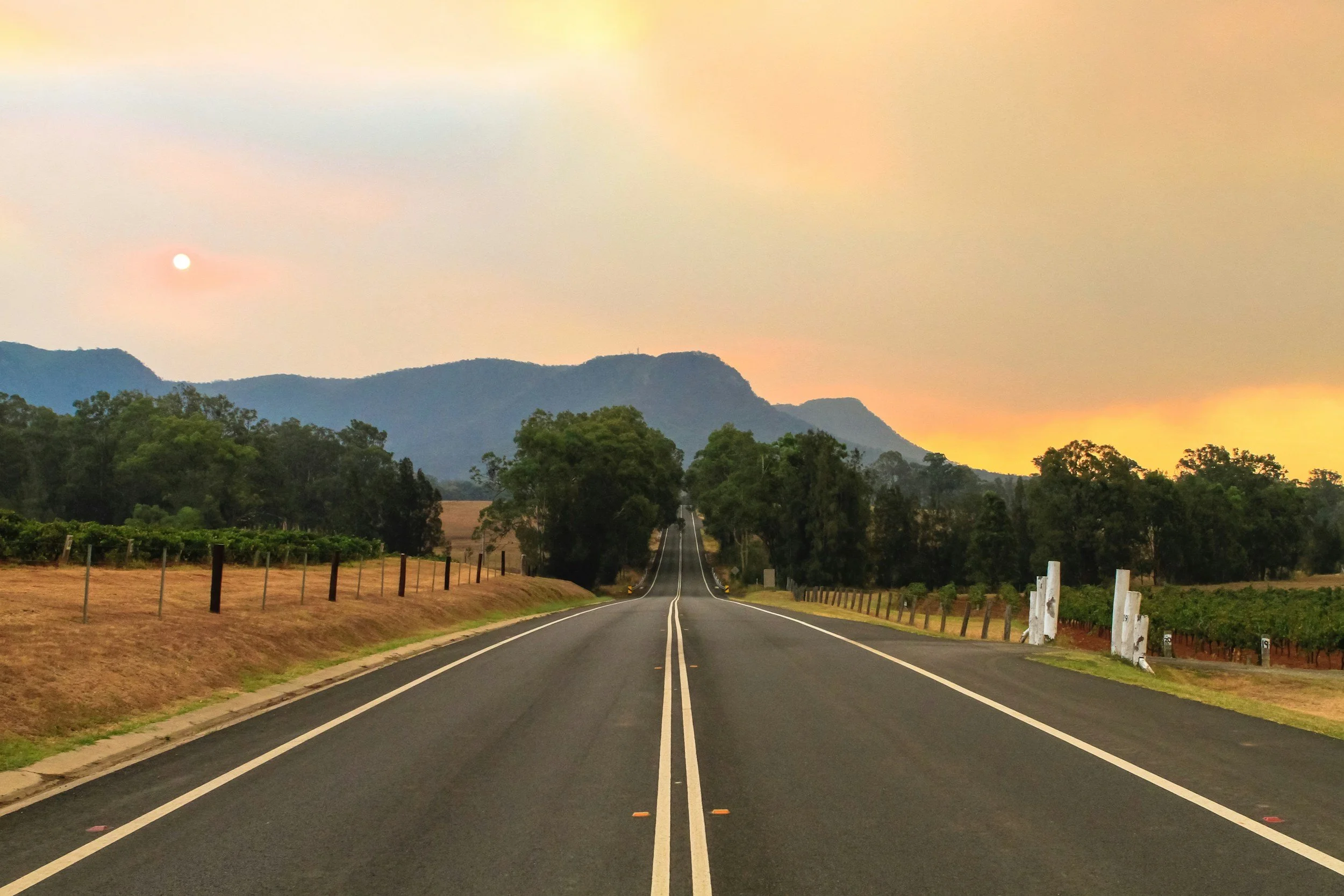 A straight two-lane road stretching into the horizon, flanked by green trees and farmland, with mountains in the background and a sunset sky.