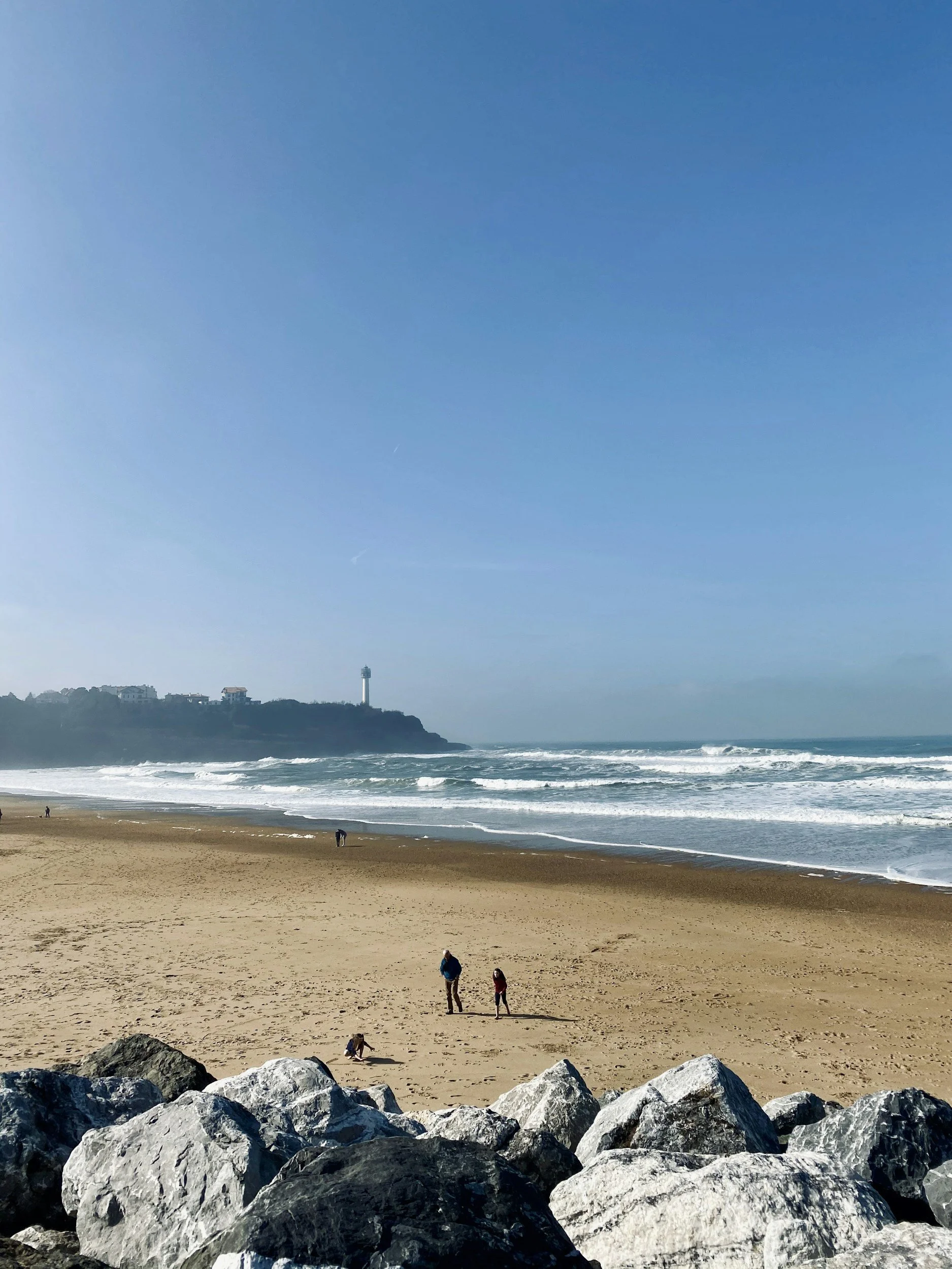 A sandy beach with large rocks in the foreground, a few people walking, and a lighthouse on a distant hill under a clear blue sky.