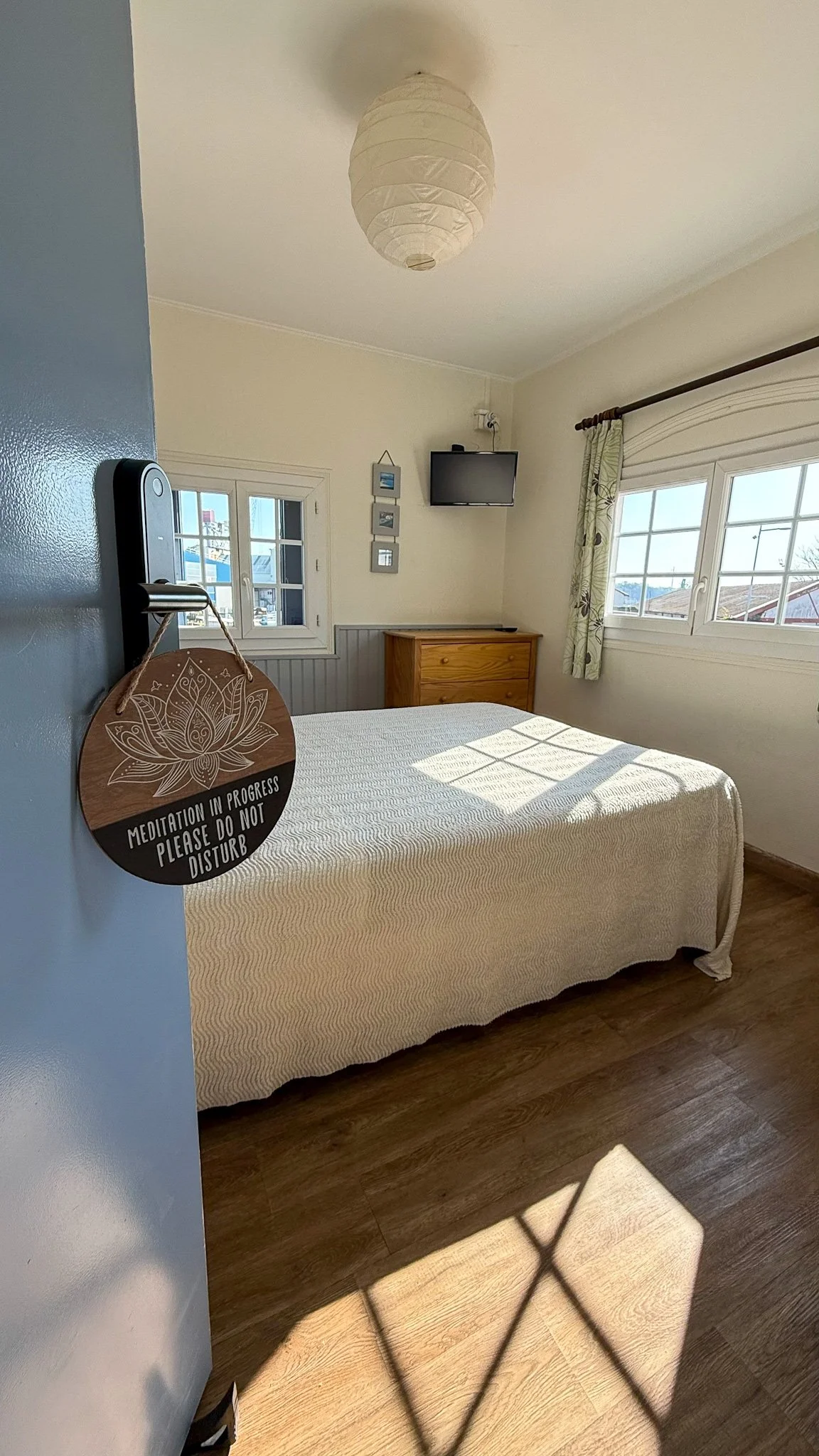 A cozy bedroom with a wooden bed frame, white bedding, and a beige throw blanket. There are two nightstands with lamps, a window with blue curtains, and wall decorations including a straw hat and framed art. A small desk with a potted plant is in the corner, and a rug covers part of the hardwood floor.