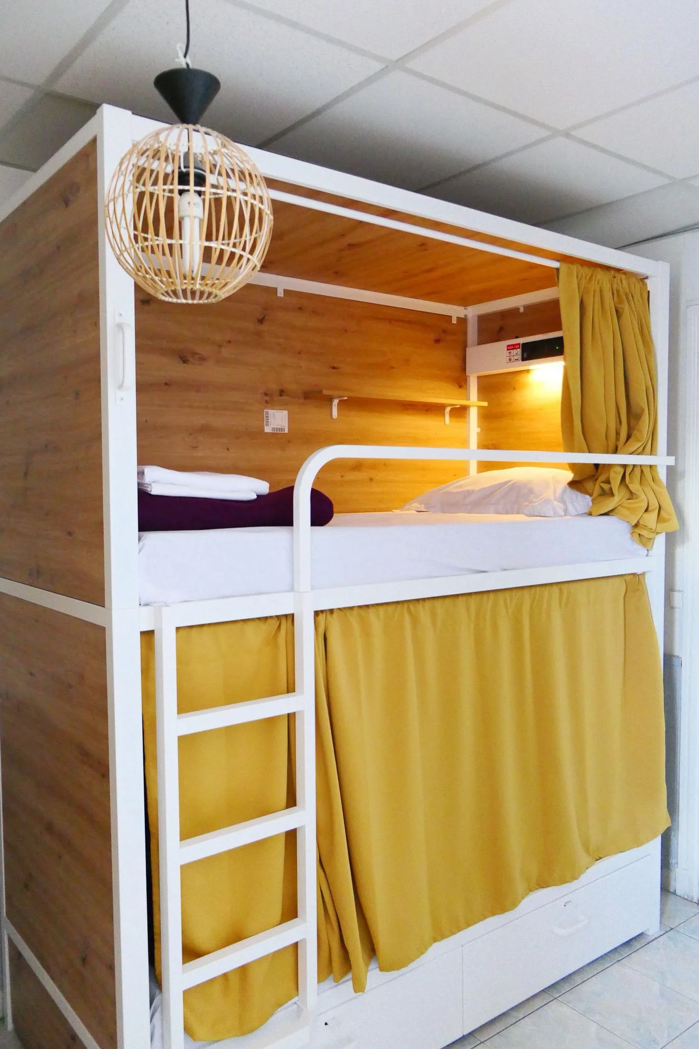 A cozy dormitory room with two sets of bunk beds featuring yellow curtains and bedding, a small round rug on tiled floor, warm lighting, and a window with yellow drapes.