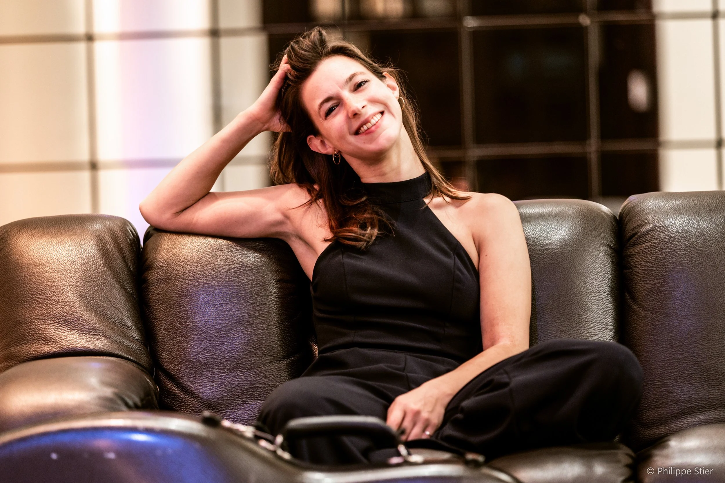 A smiling woman with brown hair sitting casually on a leather couch at home or in a lounge. She is wearing a black sleeveless top and black pants, with one arm resting behind her head. The background features a modern, geometric wooden wall. Her expression conveys happiness and relaxation.