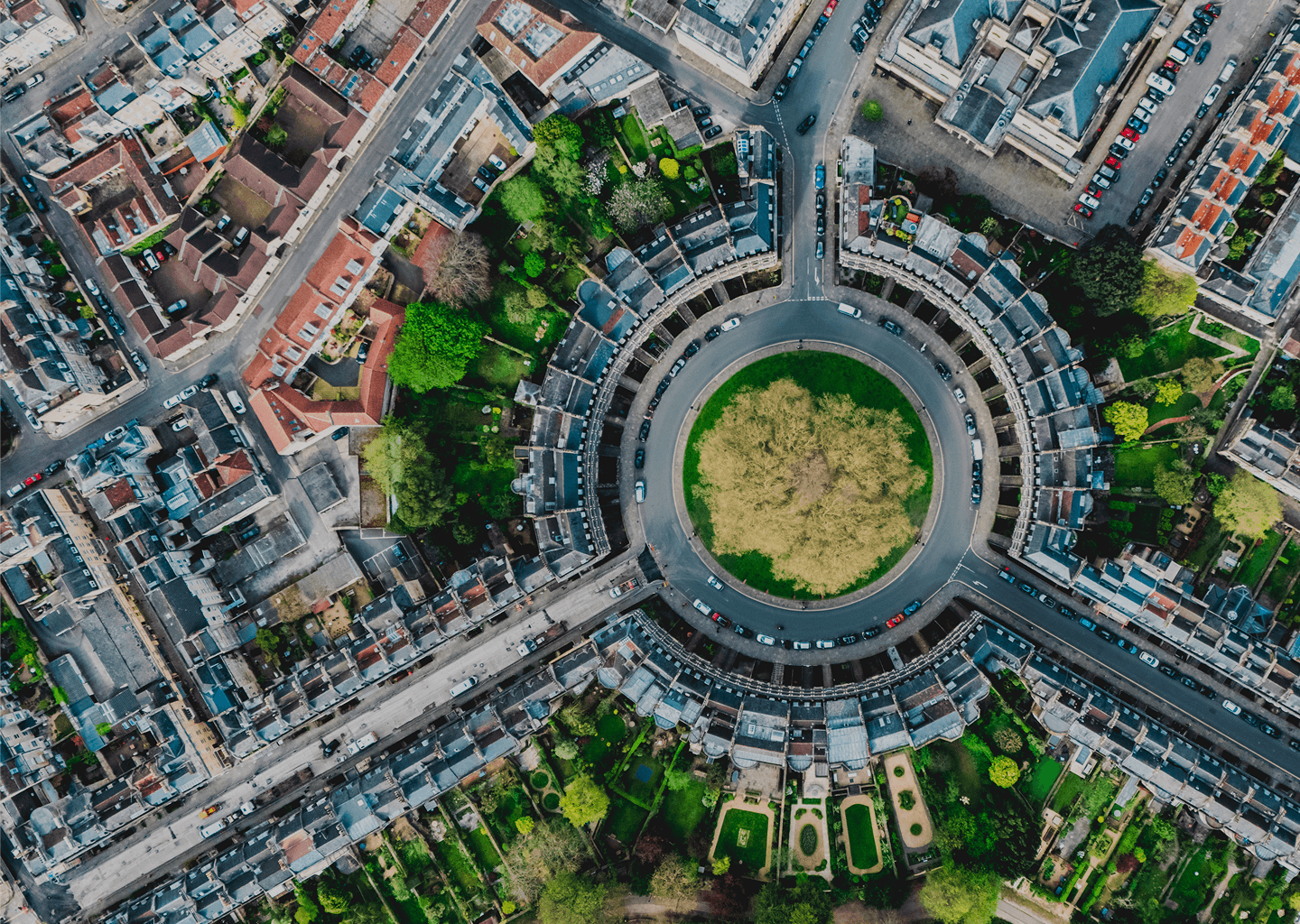 Aerial view of a city with a circular park at the center, surrounded by residential and commercial buildings with rooftops, streets, and parked cars.
