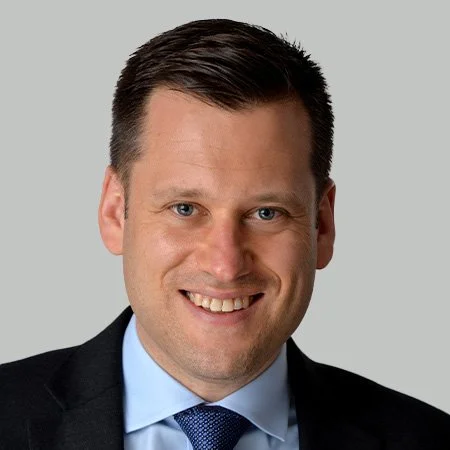 Headshot of a smiling man in a suit and tie, with short dark hair, against a light gray background.