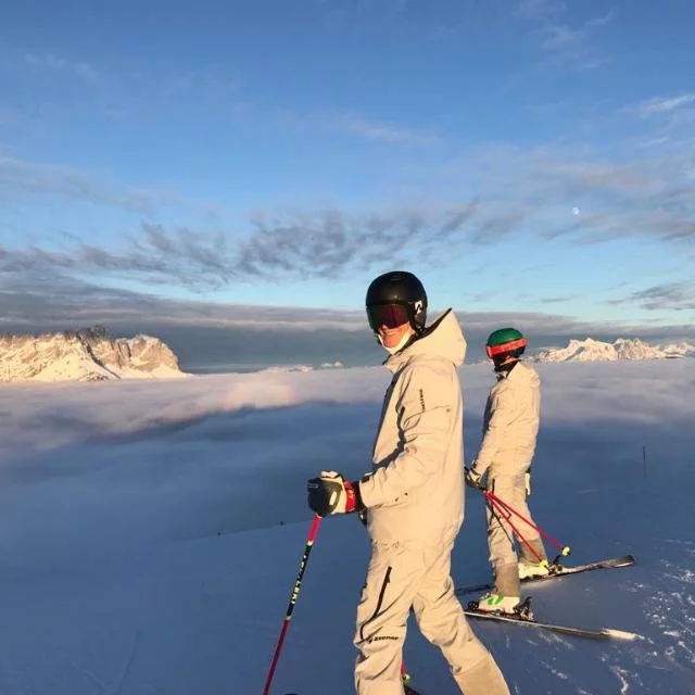 Zwei Skifahrer in weißen Anzügen und Helmen auf einem verschneiten Berggipfel bei Sonnenuntergang, mit Bergformationen im Hintergrund.
