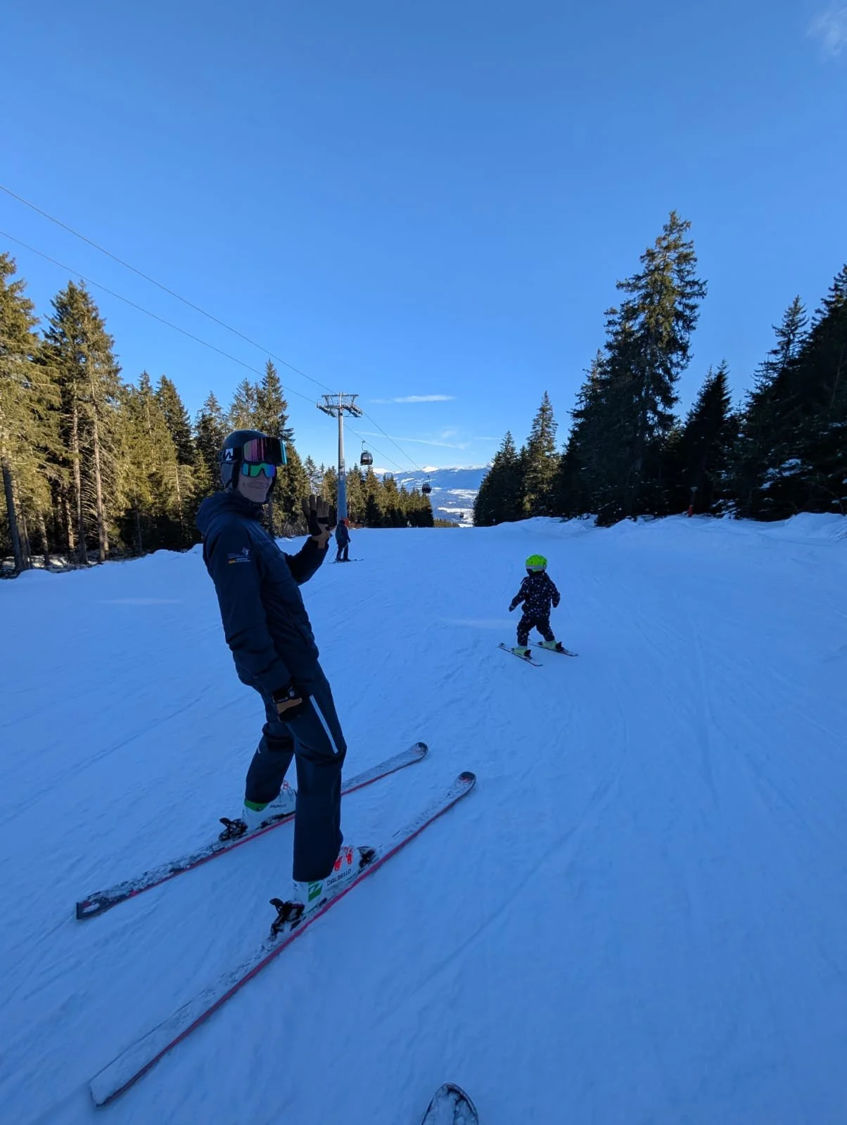 Hannes Lechner als Skilehrer mit einem Kind mit Helm, auf einer verschneiten Skipiste, im Hintergrund Tannenbäume und eine Seilbahn unter klarem blauen Himmel.