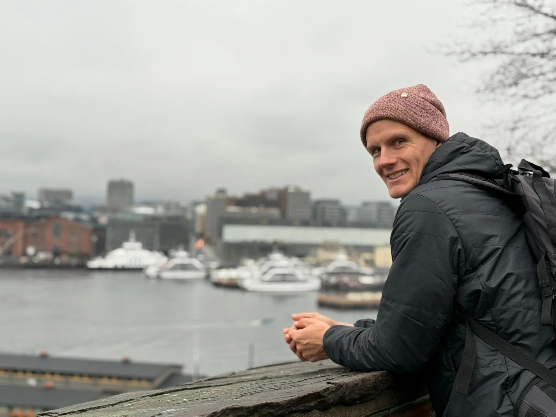 Hannes Lechner, lächelnder Mann mit Mütze und Rucksack steht an einer Brüstung mit Blick auf einen Hafen mit Yachten und Hochhäusern bei bewölktem Himmel.