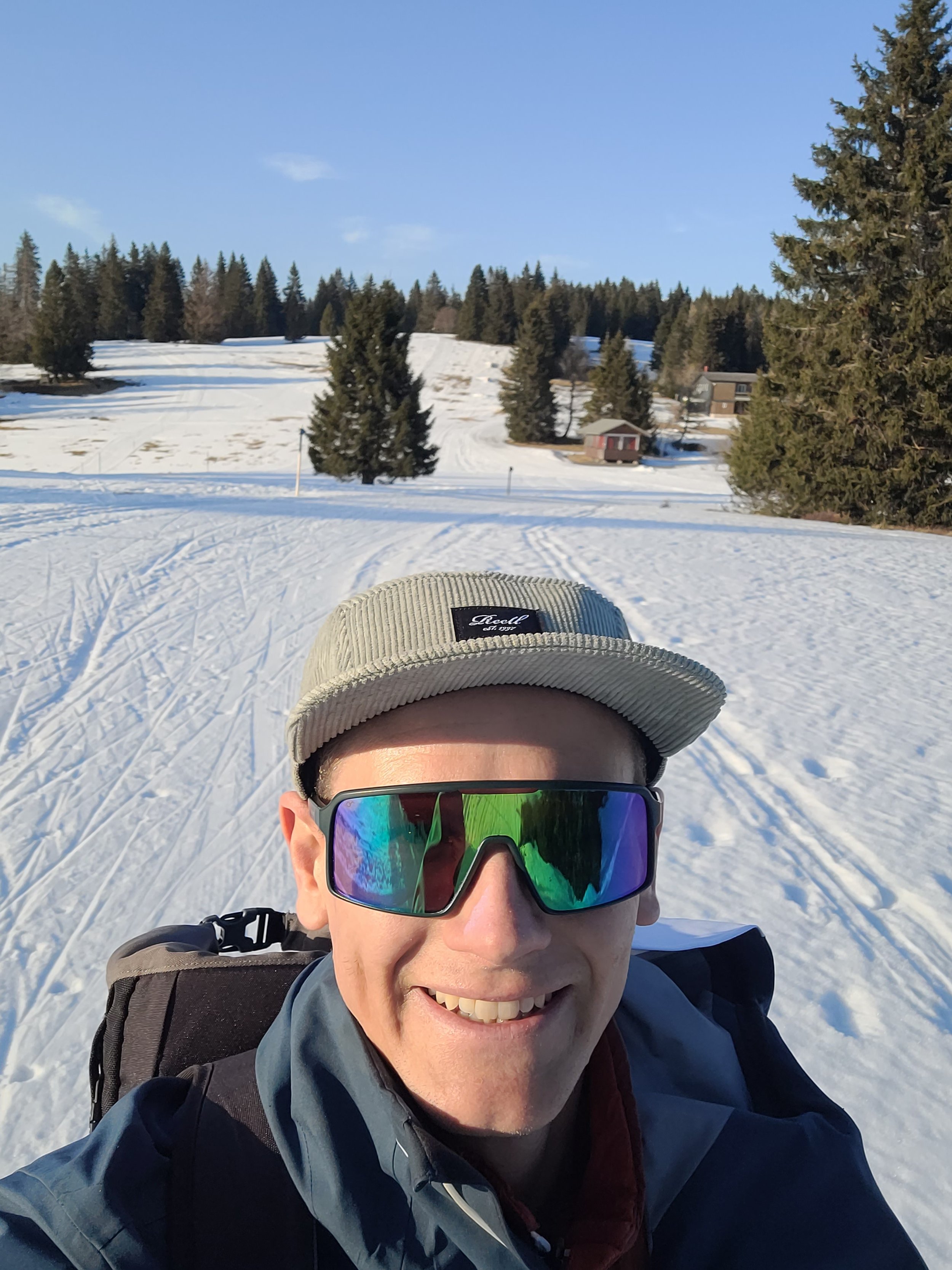 Ein Mann in Schnee mit Sonnenbrille, beige Mütze, Rucksack und Winterkleidung, im Hintergrund Berge, Tannenbäume und kleine Häuser bei sonnigem Himmel.