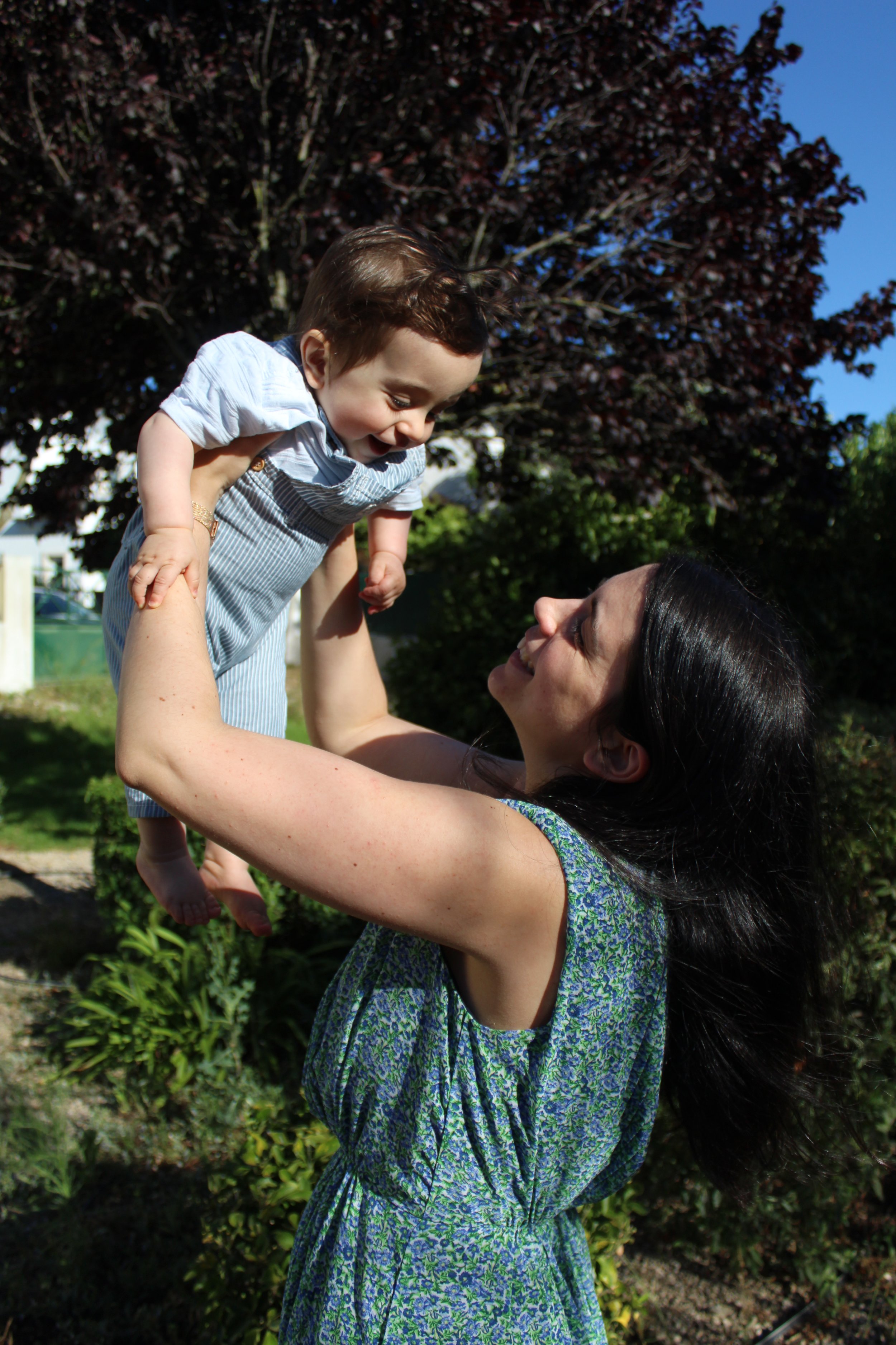 Uma mulher sorridente brincando com uma criança ao ar livre, segurando-a no ar em um dia ensolarado.