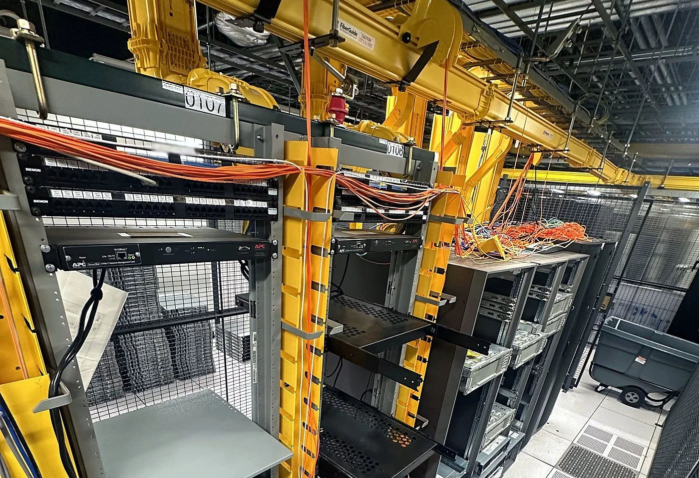 Colocation cage build-out with open racks, overhead yellow fiber raceway and orange fiber patching