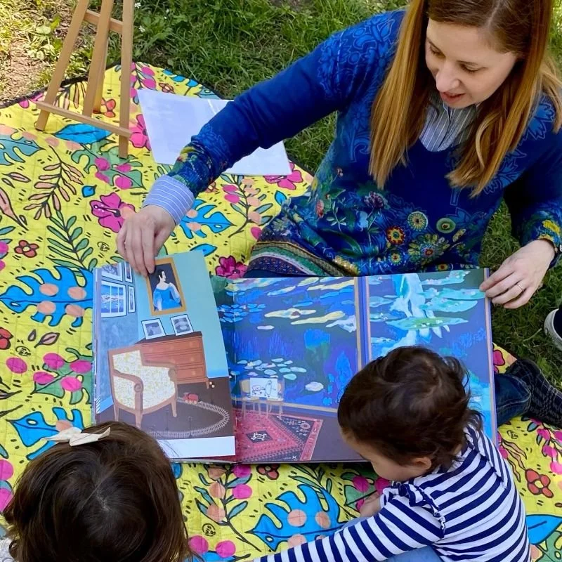 Donna che legge un libro illustrato a due bambini all'aperto su un tappeto decorato con fiori e foglie, con un piccolo cavalletto vicino.