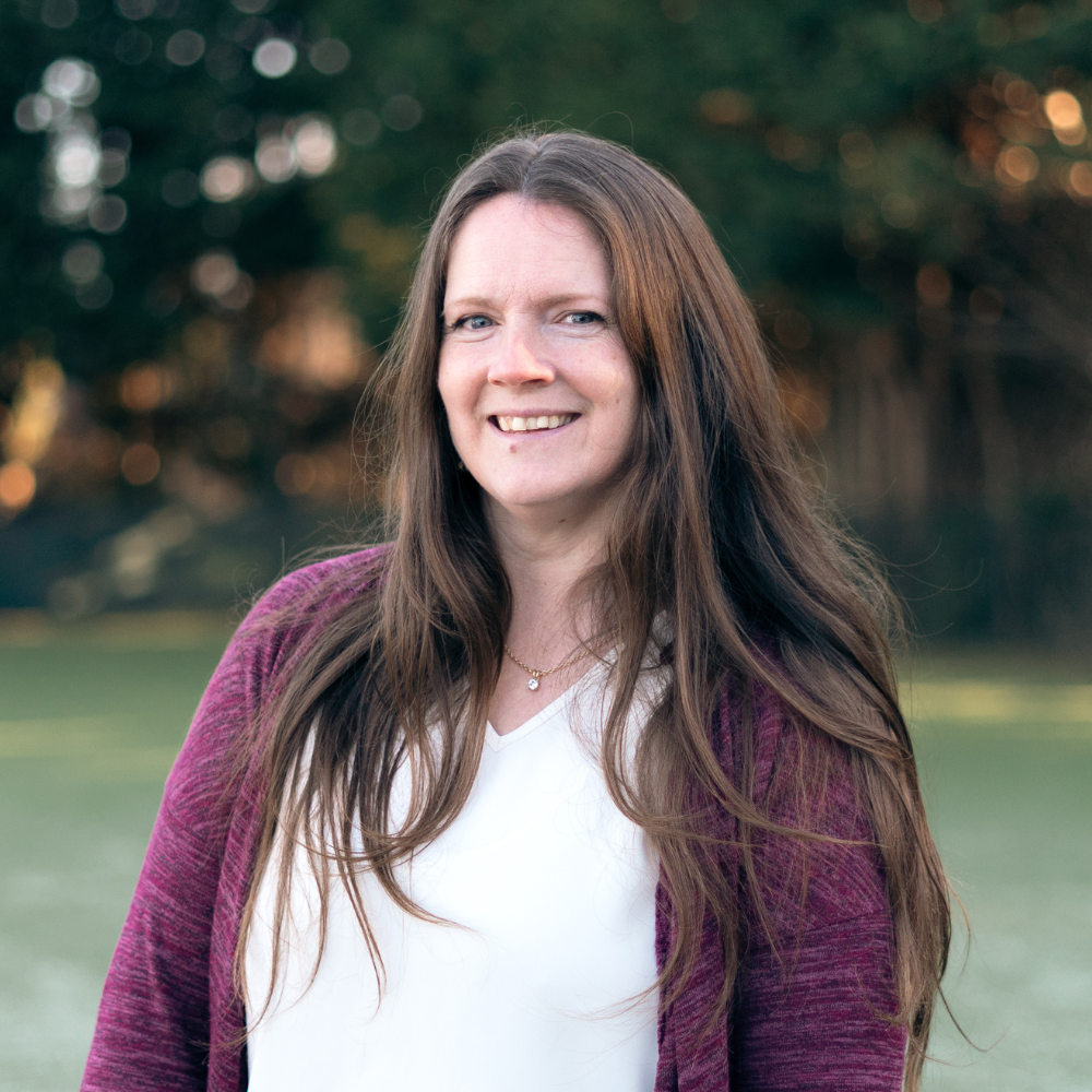 A woman with long brown hair wearing a white top and a purple cardigan, standing outdoors with trees and a blurred green background, smiling at the camera.