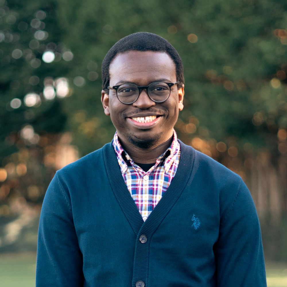 A man and woman standing close together in front of a black background, both wearing black shirts. The woman has curly hair, a necklace with a cross, and red lipstick. The man has short hair, glasses, and is smiling.