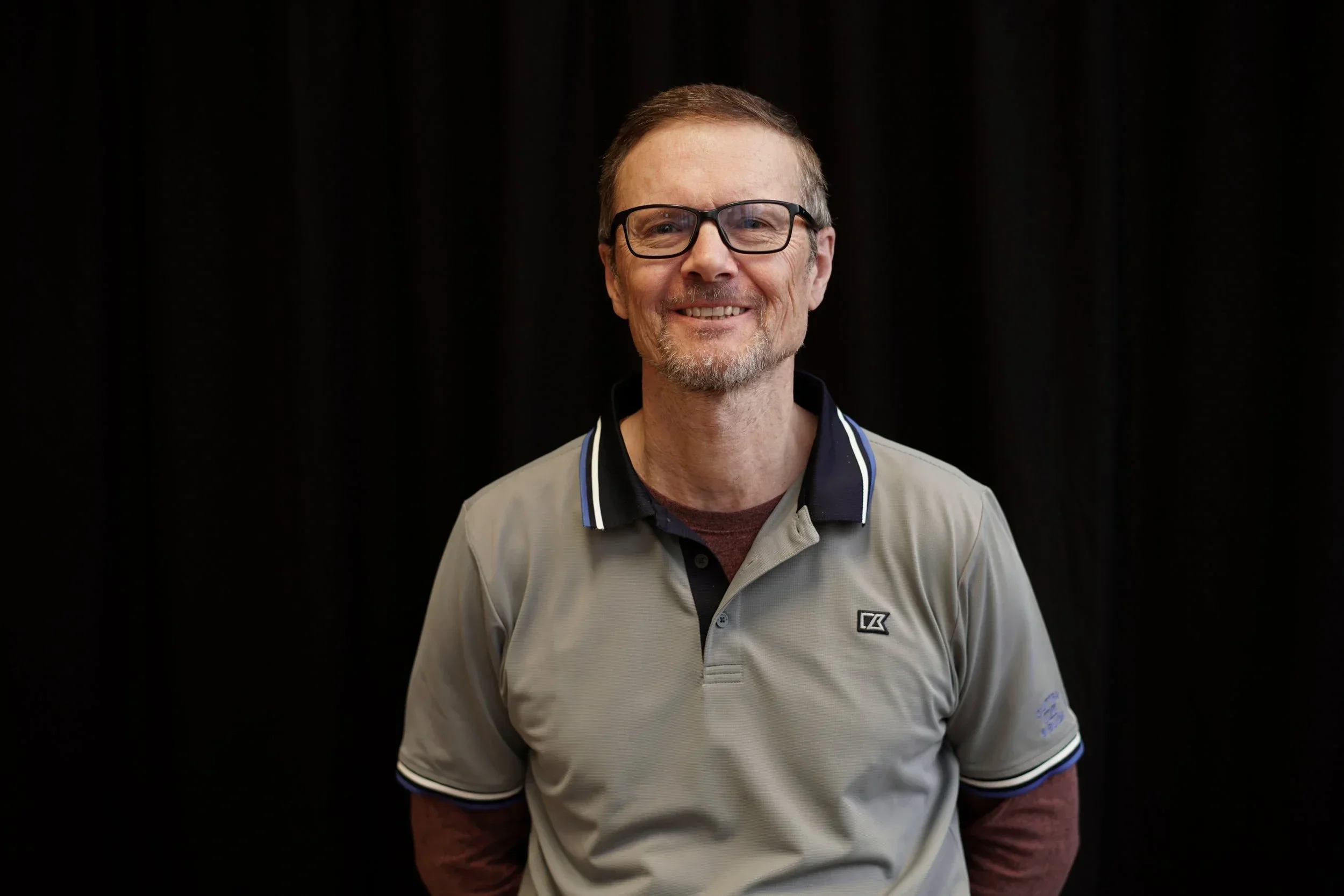 A man with glasses and a beard, smiling, wearing a light gray polo shirt with black and white accents, standing against a black background.