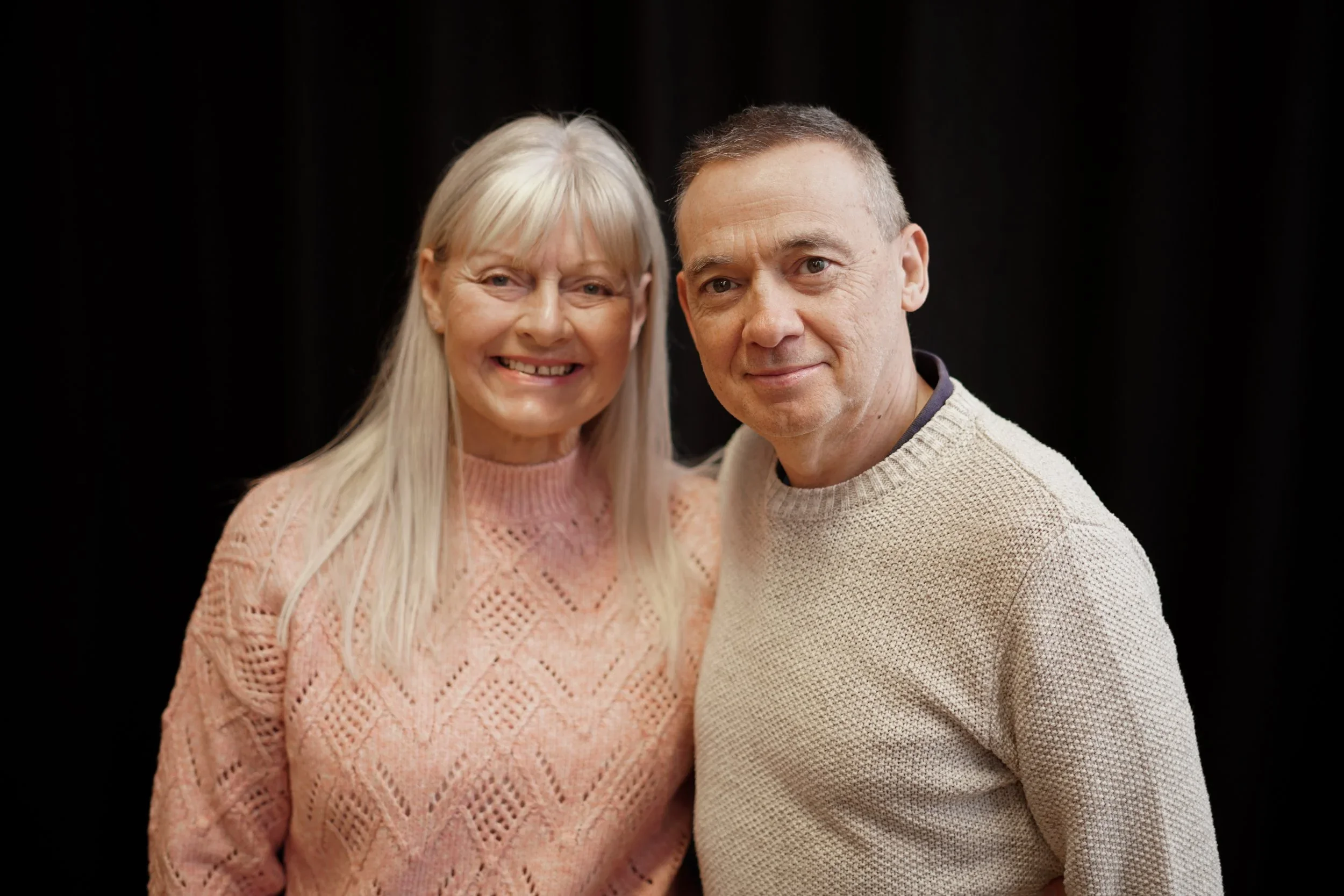 A smiling woman with long hair wearing a pink knitted sweater, standing next to a man with short hair wearing a beige sweater, both posing against a dark background.
