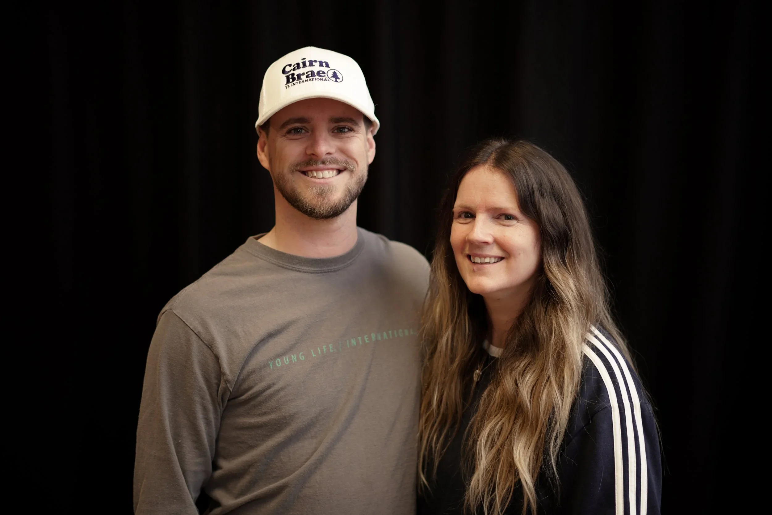 A smiling man wearing a white cap and a gray t-shirt standing next to a smiling woman with long wavy hair wearing a black jacket with white stripes on the sleeves, against a black background.