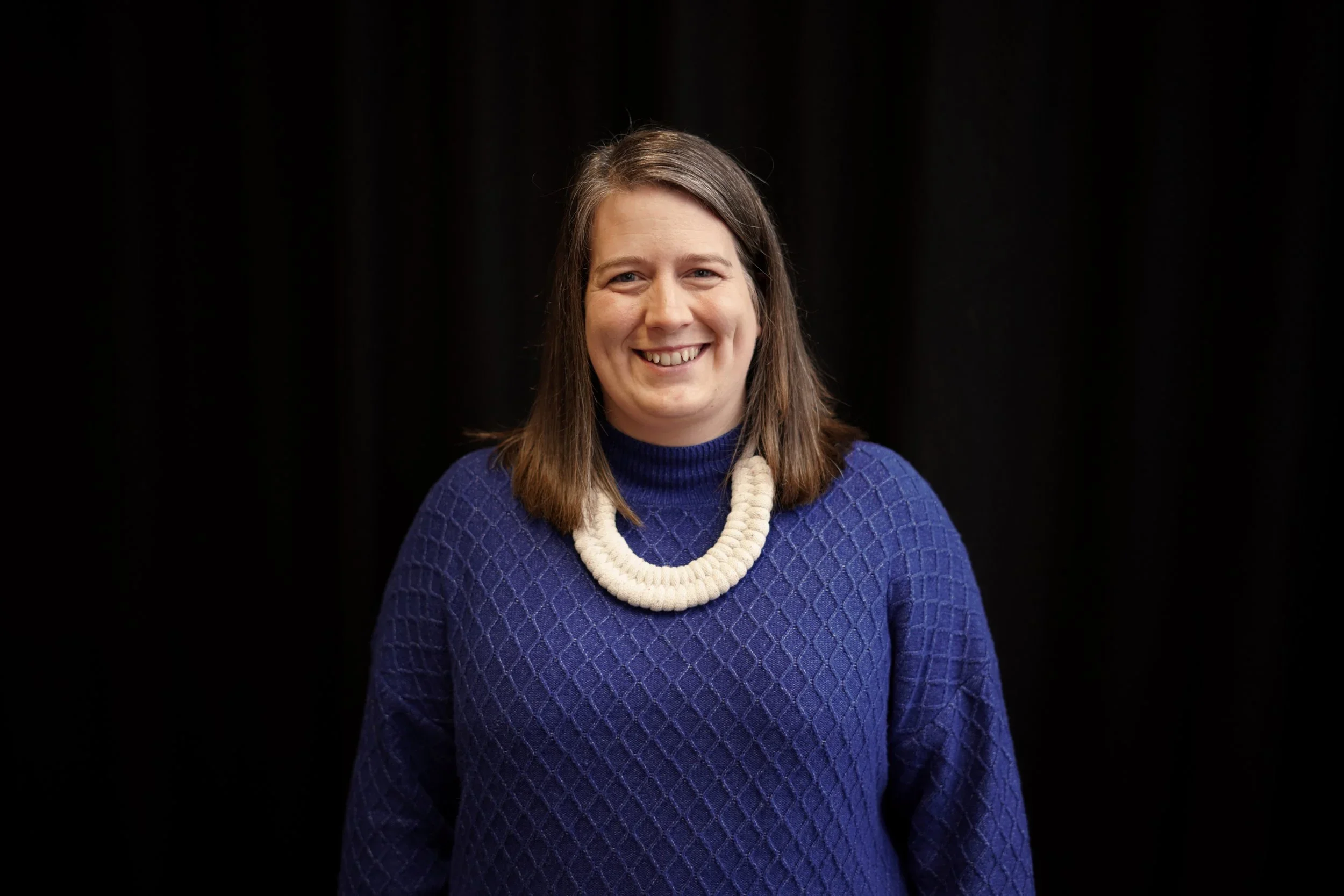 A woman with shoulder-length brown hair, wearing a blue textured sweater and a white chunky necklace, smiling at the camera against a black background.