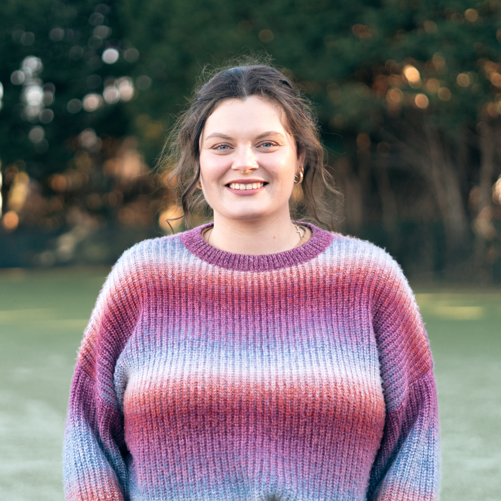 Young woman smiling outdoors in a colorful sweater, with trees in the background.