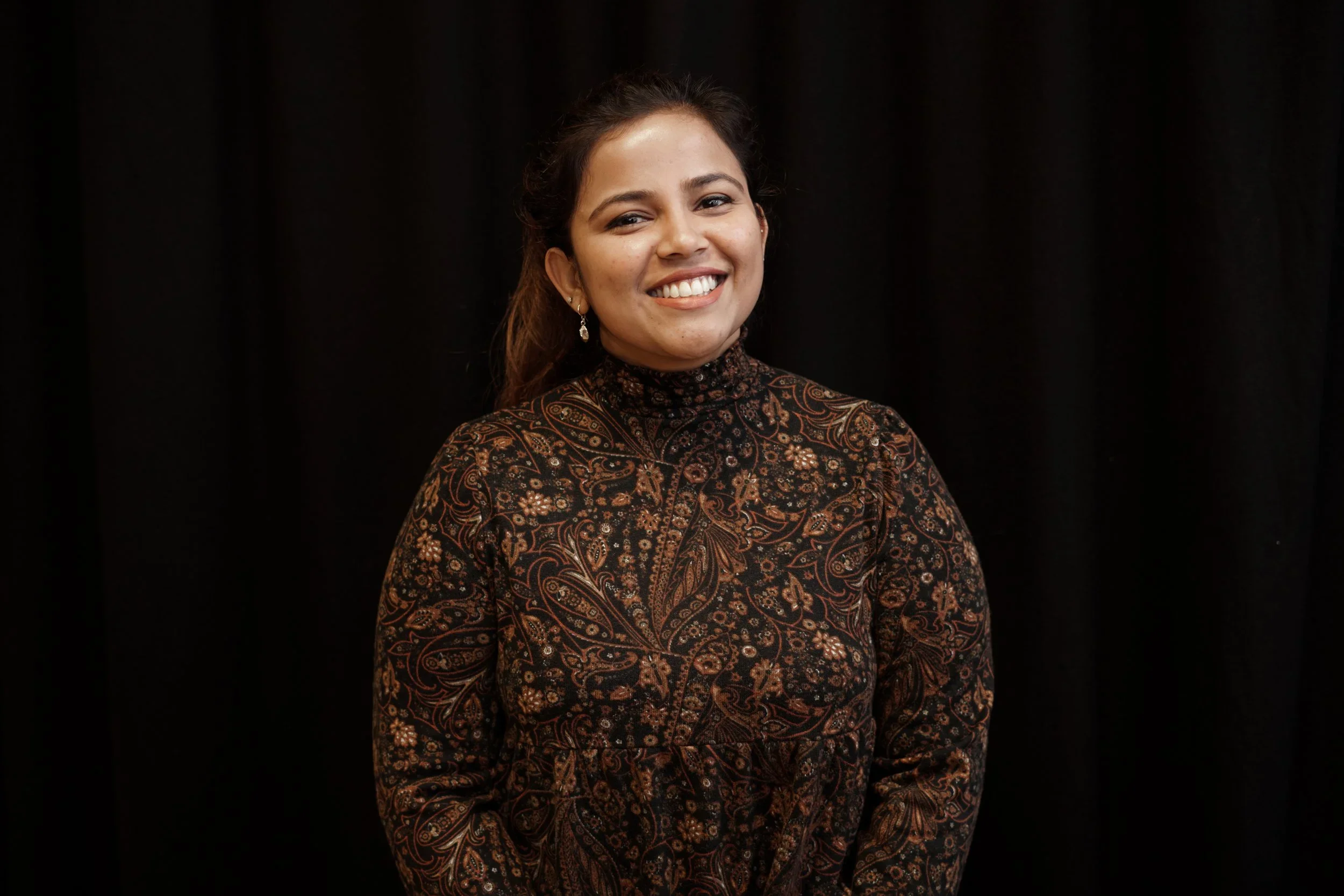 A woman smiling in front of a black backdrop, wearing a patterned high-neck dress and earrings.